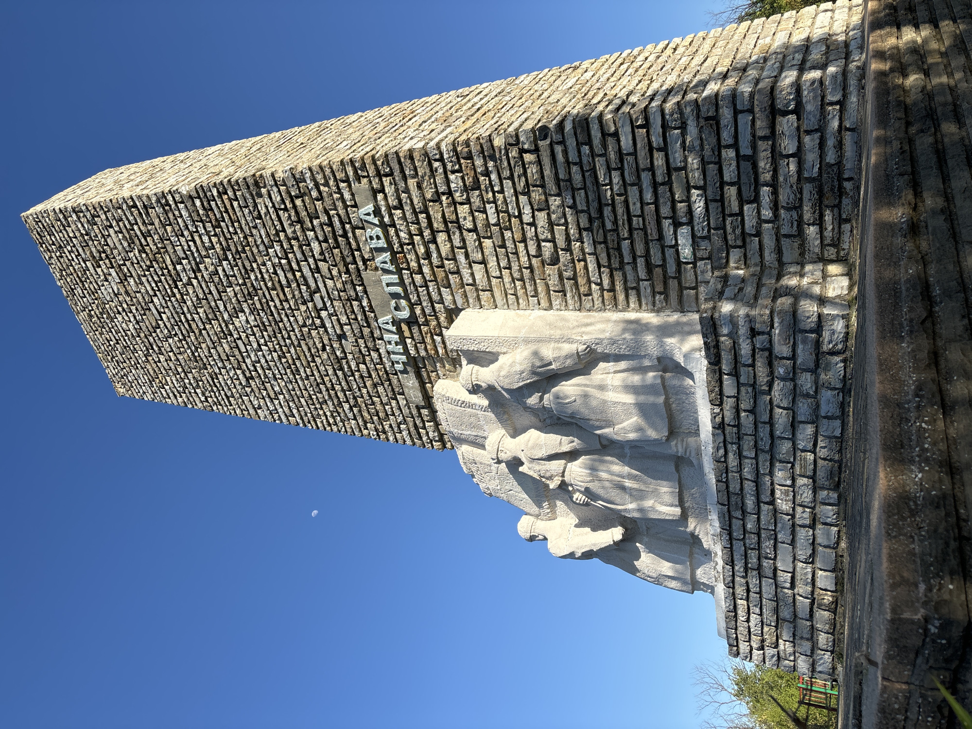 Here is a description of the image:
The image shows a monument against a clear blue sky. The monument appears to be constructed of light-colored, roughly rectangular stones stacked to form a tall, thick structure. There is a relief carving of several figures affixed to the front of the stone structure. Inscribed on the monument is text in a non-Latin alphabet. At the base, the monument transitions to darker-colored stones. Part of the landscape is visible including sparse greenery and what seems to be play equipment. A small moon is visible in the sky. The location appears to be Unknown, Unknown.
