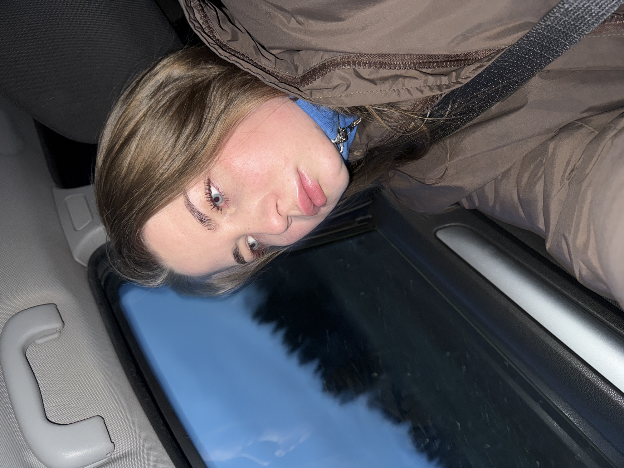 A close-up portrait features a young woman looking directly at the camera from inside a vehicle. She has light brown hair, fair skin, and striking blue eyes, with her lips slightly parted in a neutral expression. She is dressed in a brown puffer jacket, under which a blue top with a metallic zipper pull is visible. A dark gray, textured seatbelt crosses her shoulder and chest area.
The setting is clearly a car interior. To her left, a light gray fabric ceiling and a matching gray grab handle are visible. Behind her, a dark gray or black car seat or headrest can be seen. To her right, a car window reveals a reflection of a clear blue sky and dark, silhouette-like shapes, likely trees or a forest, indicating an outdoor, natural environment. Below the window, part of the car door interior is visible, featuring a silver trim accent and black textured material.
The scene is captured during the daytime, suggested by the bright blue sky reflected in the window. The woman's expression appears calm and composed, perhaps slightly inquisitive. There is no visible text present in the image. While the provided context specifies the location as Ol'khovochka, Russia, there are no distinct visual cues within the image itself to confirm this geographical detail.