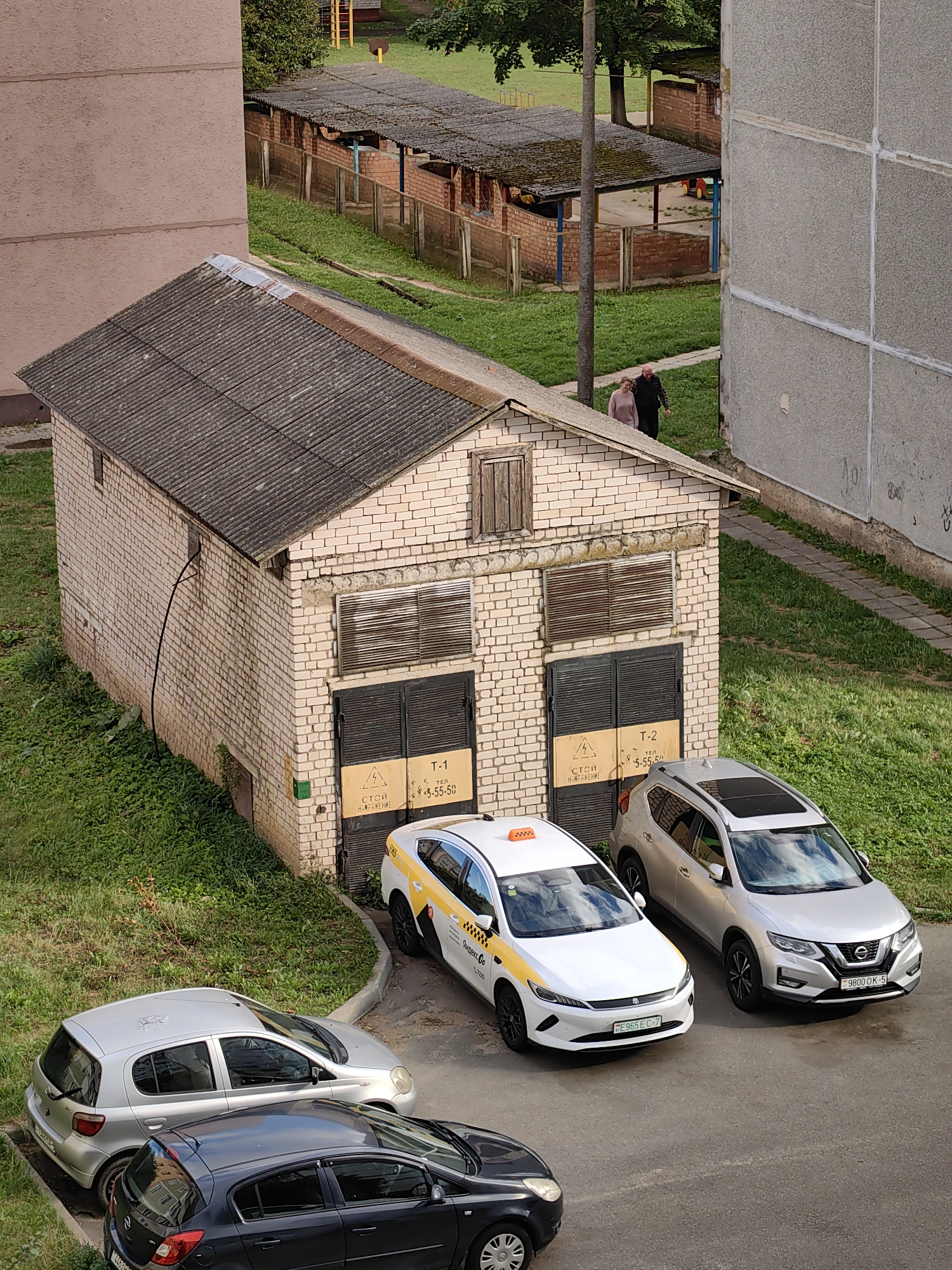 In Fanipal, Belarus, a side view captures a small, light-colored brick building with a corrugated metal roof. The building has two large dark garage doors, each with a yellow warning sign depicting a lightning bolt and text. Above the doors are horizontal slatted windows. To the right of the building, two silver cars are parked: a smaller hatchback and a larger SUV. In front of the building, a white taxi with yellow accents is parked, its roof adorned with an orange taxi sign. A dark gray hatchback is parked in the foreground, angled away from the viewer. The area surrounding the building is covered in green grass and vegetation. In the background, two people walk away from the camera along a path. Further back, a covered outdoor structure with a similar corrugated roof and brick supports is visible, along with a playground in the distance. The scene appears to be in a residential or industrial area with buildings and a yard.
