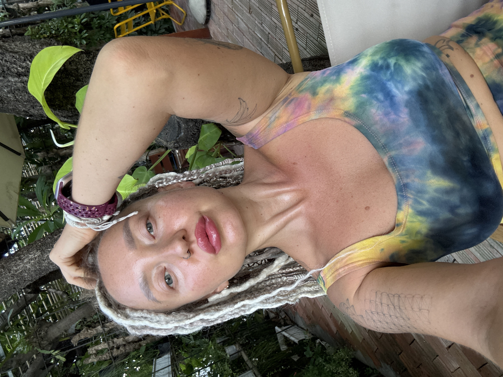 The image is a close-up, vertically oriented selfie of a person lying down outdoors. The person has fair skin, light-colored eyes, and blonde braids. They are wearing a tie-dye tank top in shades of blue, yellow, pink, and green. A purple watch with white string details is on their left wrist, and a nose ring is visible. The person's left arm is bent and resting behind their head, with their hand on their head. Their right arm is bent and positioned across their chest, partially visible.
The background consists of lush green foliage, a textured tree trunk, and a light-colored brick wall, suggesting an outdoor setting, possibly a garden or patio area. In the upper left corner, a yellow metal structure is partially visible. The lighting suggests it is daytime, with sunlight highlighting the person's skin, creating a slight sheen. The overall mood appears relaxed and casual.
No visible text is present in the image. The location context of Nha Trang, Vietnam, indicates a tropical or subtropical climate, which aligns with the presence of dense greenery.