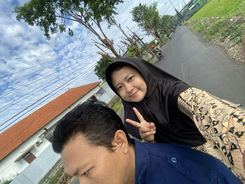 The image displays an outdoor scene on a paved road in Putat, Indonesia. In the immediate foreground, two individuals are present. A male, visible from the left side of his head and torso, wears a blue collared shirt and faces left, away from the viewer. To his right, a female wearing a black hijab and a light-colored, patterned long-sleeved garment smiles directly at the camera, extending her right hand to form a peace sign.
The background features a building with a white facade and an orange-tiled roof on the left. Numerous overhead utility lines crisscross the blue sky, which contains scattered white clouds. The asphalt road extends into the distance, bordered by trees, some with bare branches, on the right. Farther down the road, at least two motorcycles are visible, one with a rider wearing a red helmet. Green vegetation, consistent with agricultural fields, is present along the right edge of the distant road.