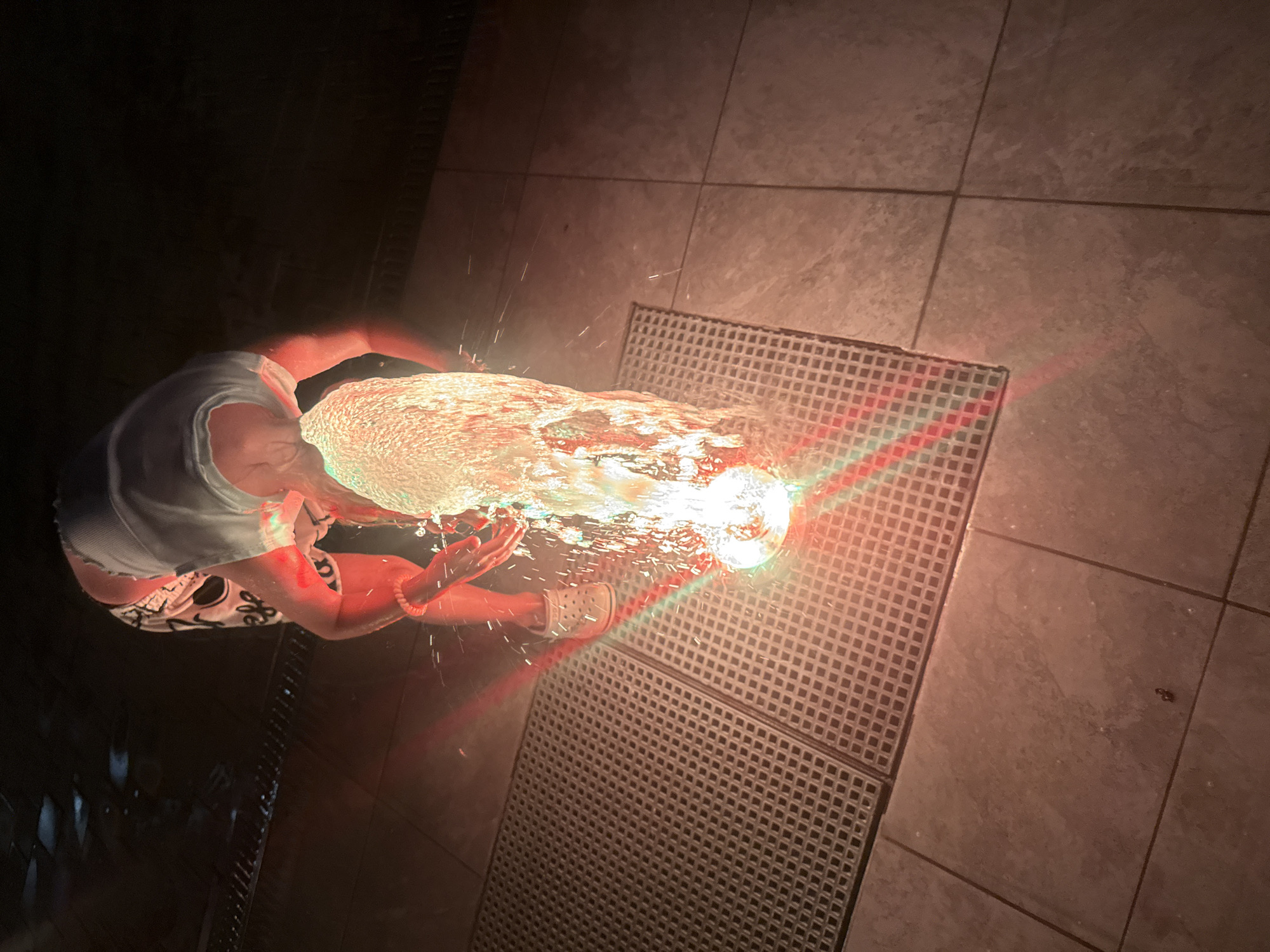 A person, viewed from above and slightly to the side, is crouched over a water feature at night. The person is wearing a light-colored t-shirt and patterned shorts, and white clogs. Their arms are submerged in the water, which is illuminated by colorful lights, casting a red and green glow. Water is splashing upwards and outwards. The person appears to be interacting with the water feature. The ground is tiled with large, square tiles, and a metal grate is visible to the left of the water feature. The scene is dark, suggesting it is nighttime. The location is identified as Partenit, Ukraine.