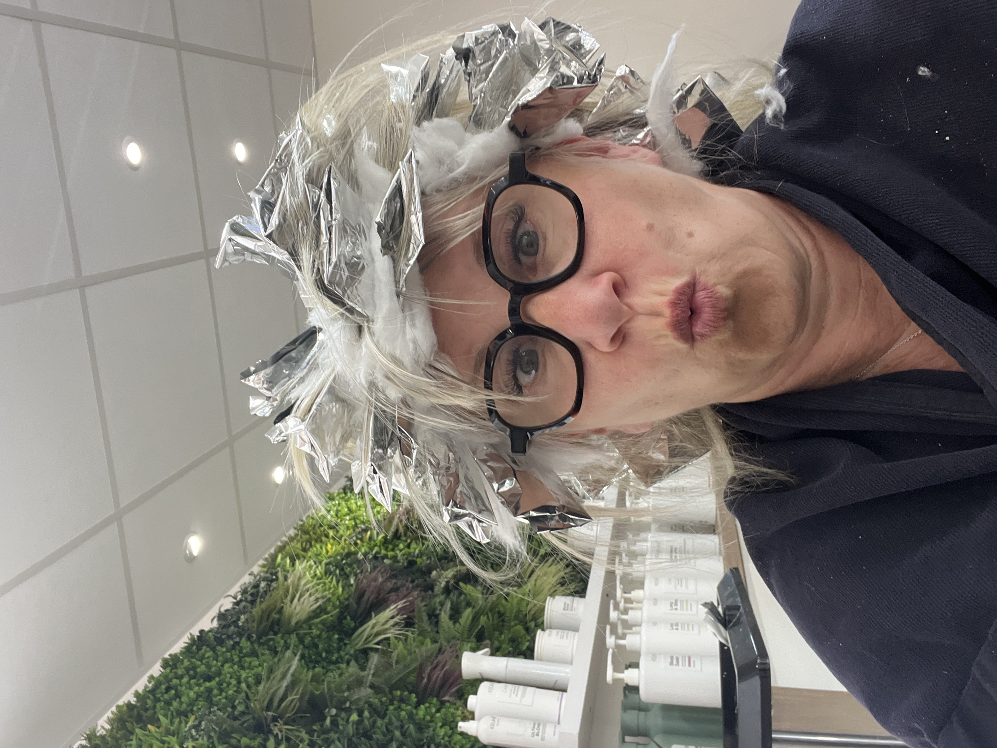 A selfie taken inside a hair salon in Montrond-les-Bains, France shows a woman with foil and cotton in her hair as part of a hair coloring process. She is wearing black-rimmed glasses and has her lips pursed. The background includes a wall with artificial greenery, bottles of hair care products, and a white tiled ceiling with recessed lights.