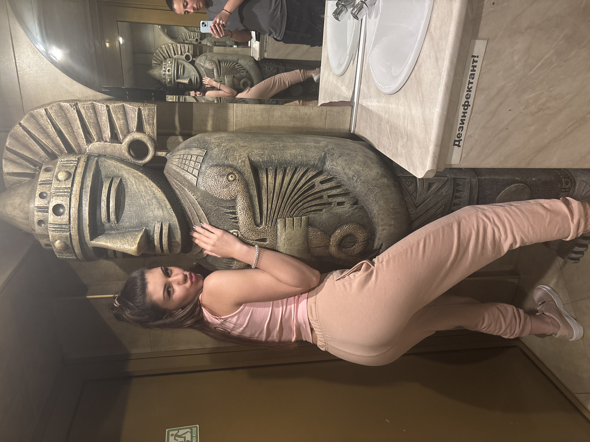 A woman in a pink tank top and sweatpants lies next to a large, ornate, stone-like sculpture of a head and torso. The sculpture appears to be a decorative element in a public restroom or similar facility, as there is a sink with dual faucets visible above it, as well as a mirror showing the scene reflected. A person holding a phone to take a picture is reflected in the mirror. The woman is holding the sculpture and looking at the camera. A sign in Cyrillic letters is on the countertop next to the sink. The setting appears to be the restroom of the restaurant "Cosmos" in Sofia, Bulgaria.