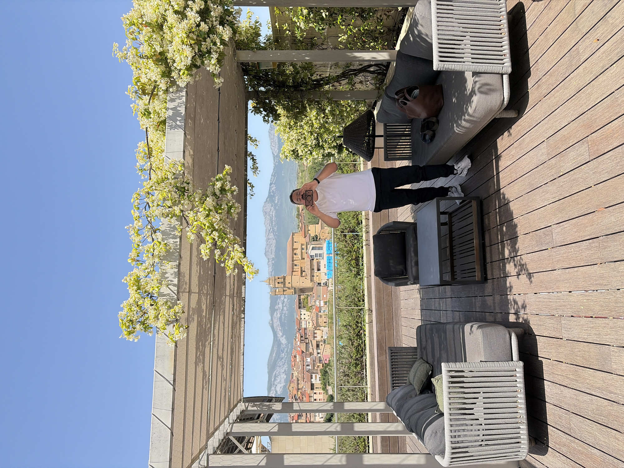 A man stands on a wooden deck, holding a phone in front of his face as if taking a picture. He wears a white t-shirt, black pants, and white sneakers. The deck is furnished with gray outdoor furniture. A structure overhead is covered in flowering vines. In the background, a cityscape is visible, with a prominent church or cathedral. The sky is clear and blue. The location appears to be a rooftop or high-level terrace overlooking the city.