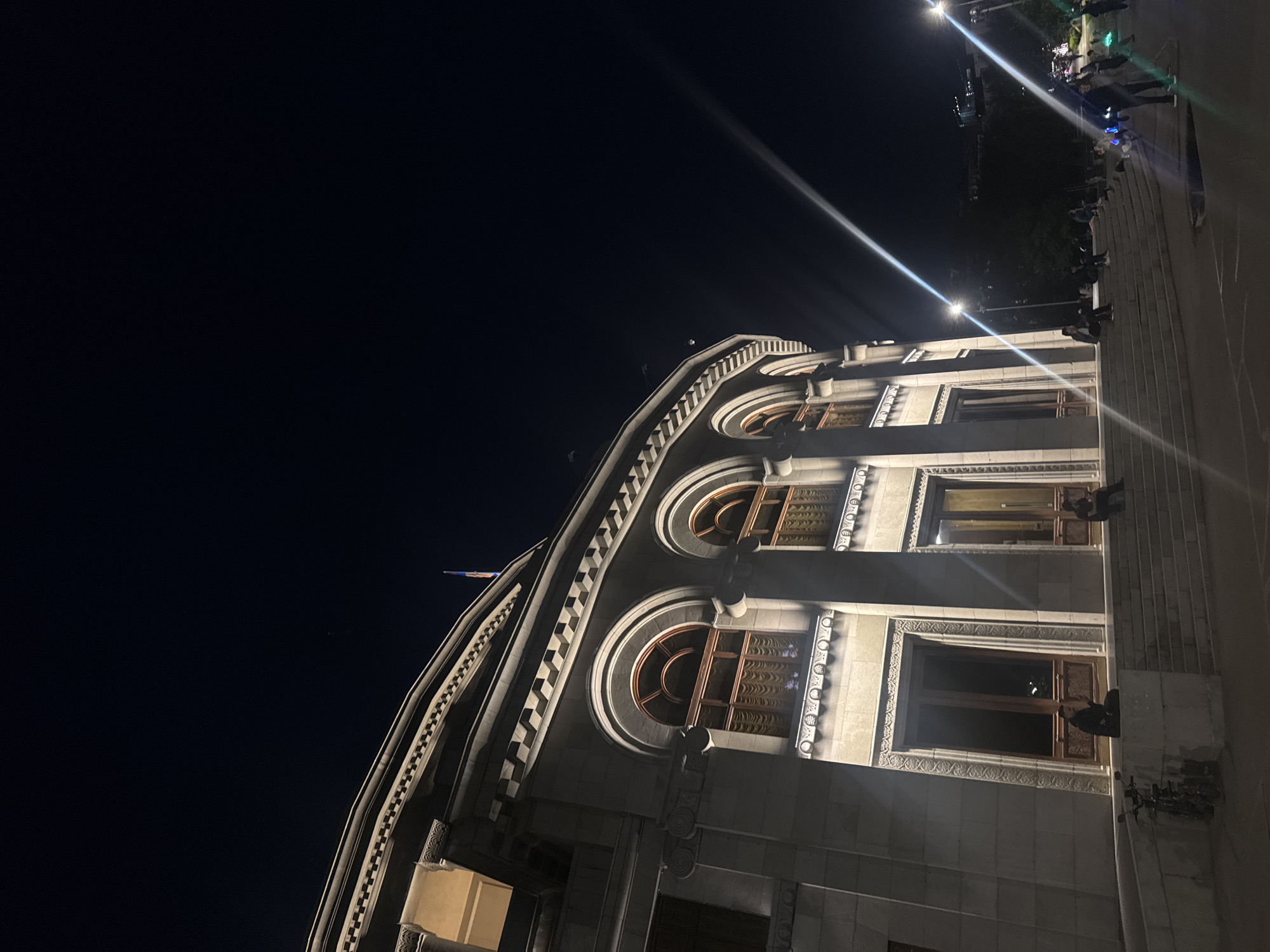 The image displays a grand, ornate building at night, photographed from a low angle with a wide, dark sky dominating the upper left. The structure, likely crafted from light-colored stone or concrete, features a distinctive curved facade adorned with intricate architectural details. Large, arched windows with decorative frames and what appear to be patterned grilles are visible across several stories. Ornate carvings, cornices, and decorative balustrades enhance the building's classical or neo-classical style. A flag is partially visible flying from the rooftop on the far left of the building.
The scene is illuminated by strong artificial lights, creating prominent lens flares that streak across the dark night sky. These lights highlight the building's texture and architectural features, casting both bright illumination and deep shadows. To the right of the building, a series of wide, ascending steps or a terraced platform is visible, where several people are gathered. Some individuals appear to be sitting or standing on these steps, while others are closer to the building's lower levels, perhaps on balconies or ledges. Their figures are silhouetted or partially lit by the surrounding lights. The overall atmosphere suggests an urban setting at night, possibly a public square or a historically significant landmark in Yerevan, Armenia. No legible text is visible in the image.