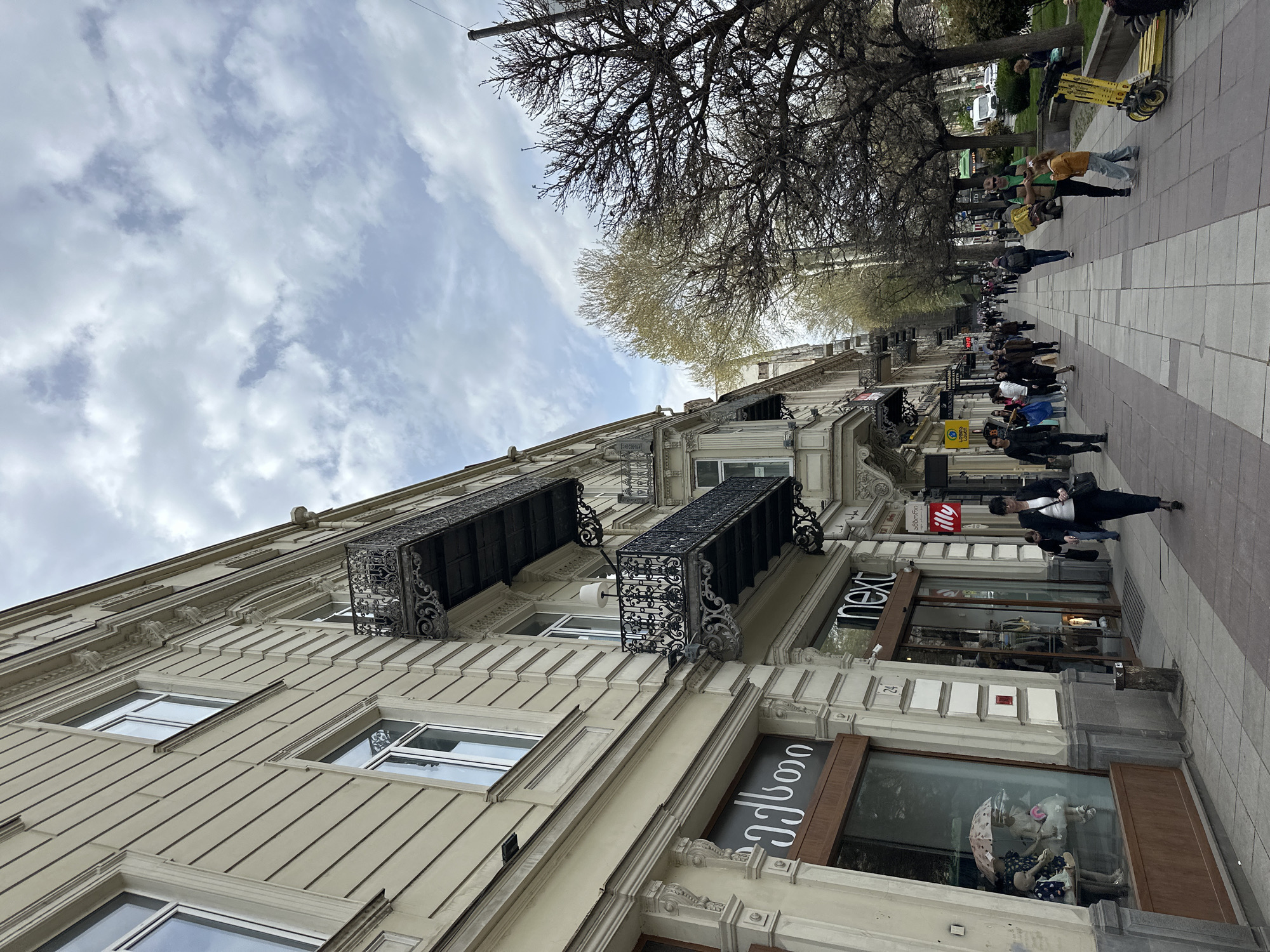The image shows a street scene in Tbilisi, Georgia. A multi-story building occupies the left side of the frame, displaying architectural details such as windows, balconies with decorative ironwork, and storefronts on the ground level. Stores like "Next" are visible. The sidewalk to the right is occupied by pedestrians walking along the street. Bare trees stand in the background, beneath a sky filled with clouds. A row of yellow scooters is parked alongside the sidewalk further down the street. The image appears to have been taken from a low-angle perspective.