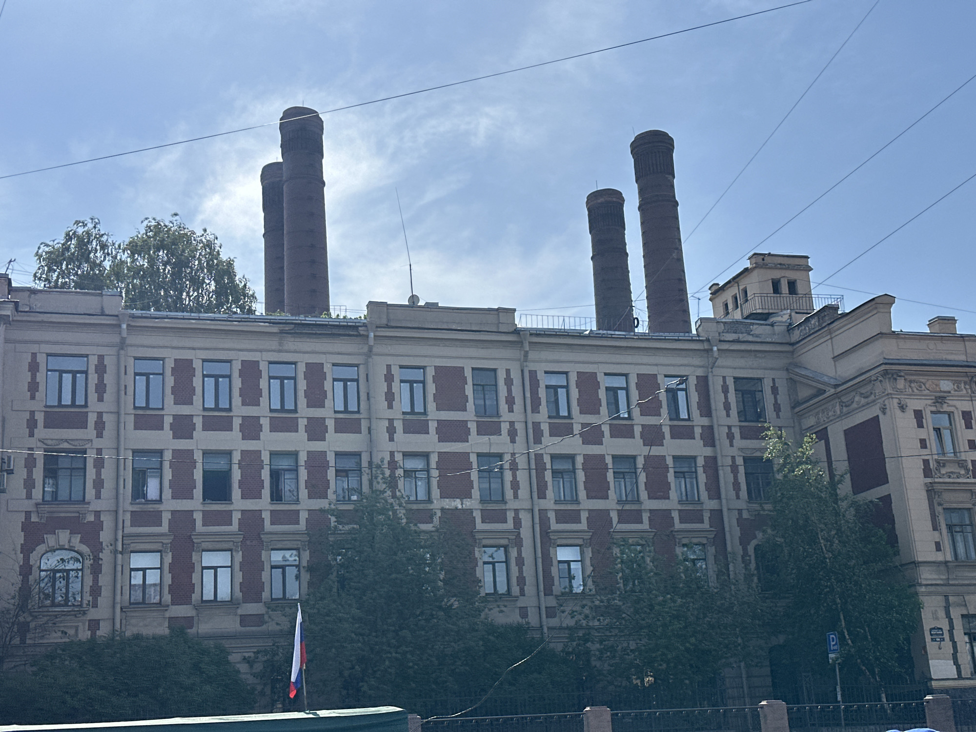 The image captures a view of the Red Banner Factory building located in Saint Petersburg, Russia. The facade of the building is visible, featuring a red-brick pattern against a lighter-colored background. The building is a multi-story structure with numerous windows. On the roof, there are tall, cylindrical brick chimneys. In the foreground, there is a glimpse of a Russian flag and some greenery. Power lines run across the sky in front of the building.