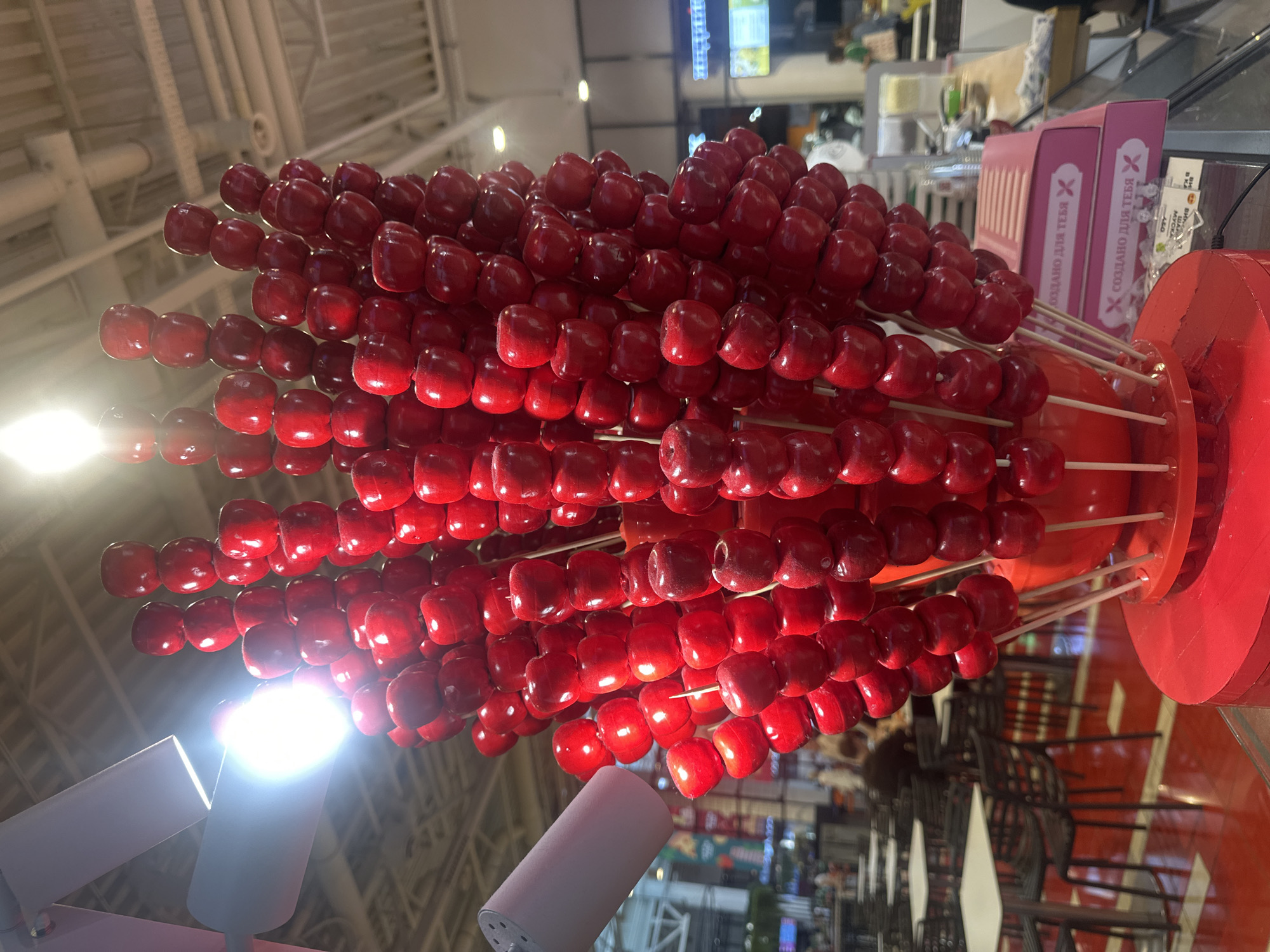 The image shows a display of candied red apples at a food stall located in the city of Khimki, Russia. The apples are skewered on sticks and arranged in a tall, vertical display, resembling a bouquet or a hand with fingers. The display is primarily red, with the apples being the dominant color. In the background, there are tables and chairs, suggesting a dining or seating area. There are also lights visible, likely overhead lighting in the establishment. The overall impression is that of a food vendor at a market or a shopping mall.