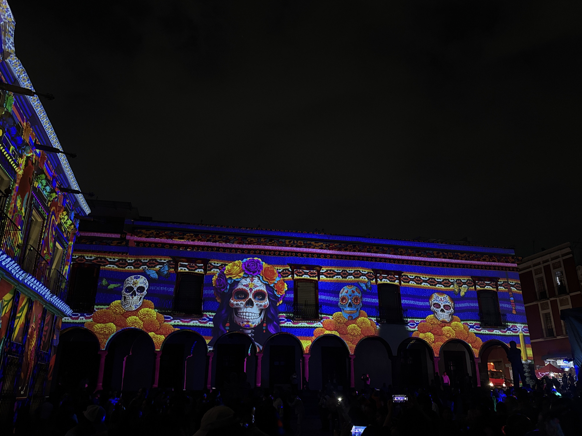 This image captures a night-time scene in the city of Puebla, Mexico, where a large crowd of people has gathered in a plaza to observe a building illuminated with projections of Day of the Dead imagery. The projections feature colorful depictions of skulls, flowers, and traditional Mexican motifs, transforming the building's facade into a vibrant display of cultural celebration. People in the crowd are using their mobile phones to capture photos and videos of the illuminated building. The atmosphere suggests a festive and commemorative event.