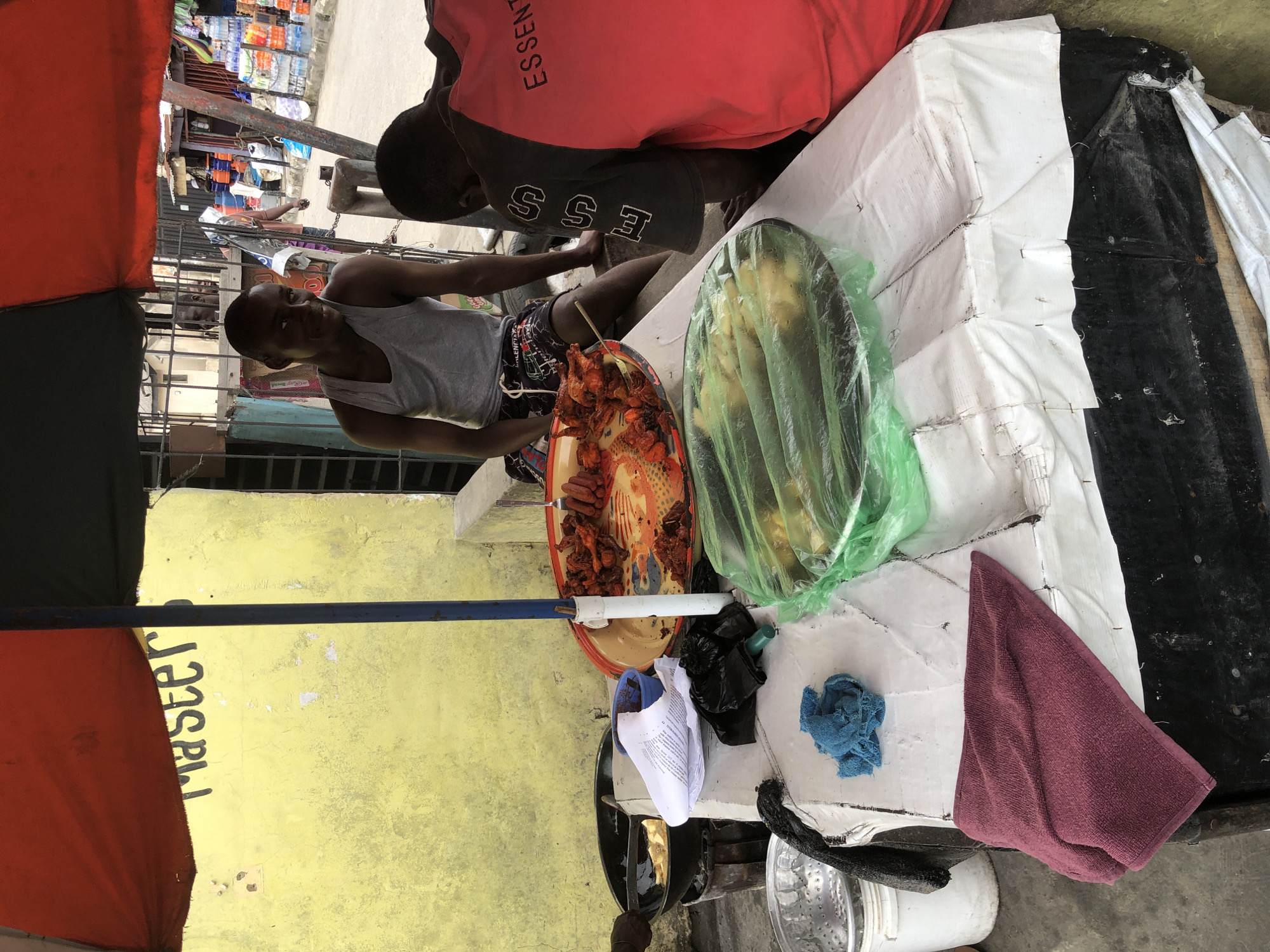 This media file depicts a scene in Ado, Nigeria, likely during the daytime under natural light, suggesting fair weather. The primary subjects are two individuals, appearing to be young men, engaged in some form of commerce or food preparation at a street-side stall. One man, wearing a grey tank top and patterned shorts, is positioned in the foreground, looking towards the camera with a neutral expression. He appears to be attending to a tray of fried food items.
Behind him, another individual is partially visible, wearing a red shirt with "ESSENTIAL" printed in black on the back, and a black garment underneath with "ESS" printed on it. They are leaning over a table covered with white and black sheeting.
On the table, several objects are laid out: a large tray holding a variety of fried foods, possibly chicken or seafood, a green plastic bag containing what appears to be more food items, and several other miscellaneous objects including a blue cloth, a white folded paper, and a maroon towel. A blue pole is visible in the foreground, supporting an orange and black umbrella that partially shades the scene. The background shows a yellow-painted wall with the word "Master" stenciled vertically in black. Beyond that, shelves stocked with various bottled goods are visible, suggesting a market or shop setting. The overall impression is of a bustling street environment where food is being prepared and sold.