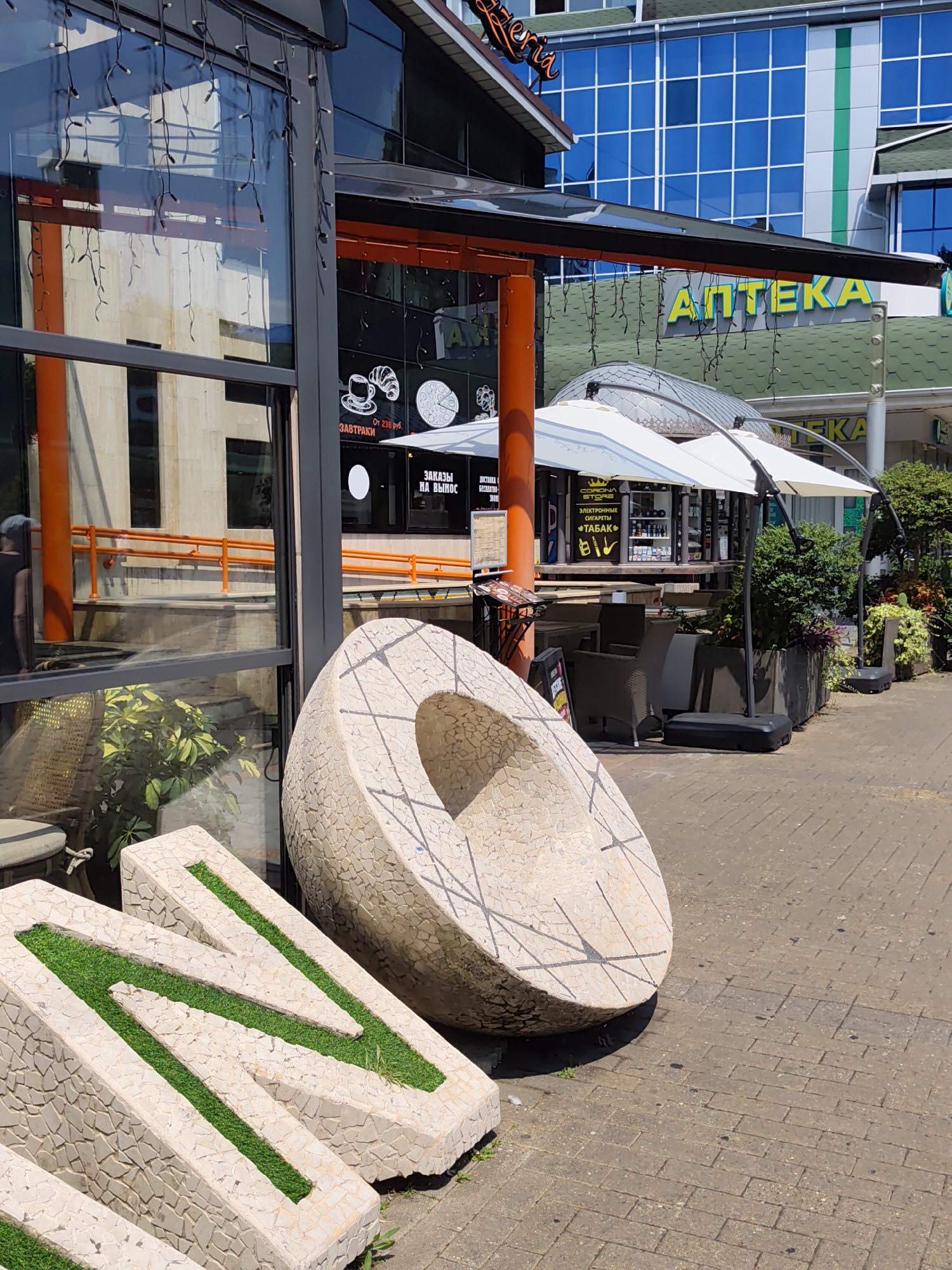 In Sochi, Russia, a sunny outdoor scene depicts a street-level view of a shopping arcade. On the left, a building with large glass windows reflects the surroundings. Decorative string lights hang from the eaves. In the foreground, two light-colored, mosaic-covered, angular planters with artificial grass are visible. Behind them, a large, round, mosaic-covered stone sculpture with carved lines is positioned on the cobblestone sidewalk. Beyond the sculpture, an outdoor seating area with tables and chairs is set up under white awnings. A sign for "АПТЕКА" (Pharmacy) with yellow lettering is prominent on a green awning in the background. Further back, a modern building with blue windows and a green stripe is visible. The overall environment is urban and commercial.