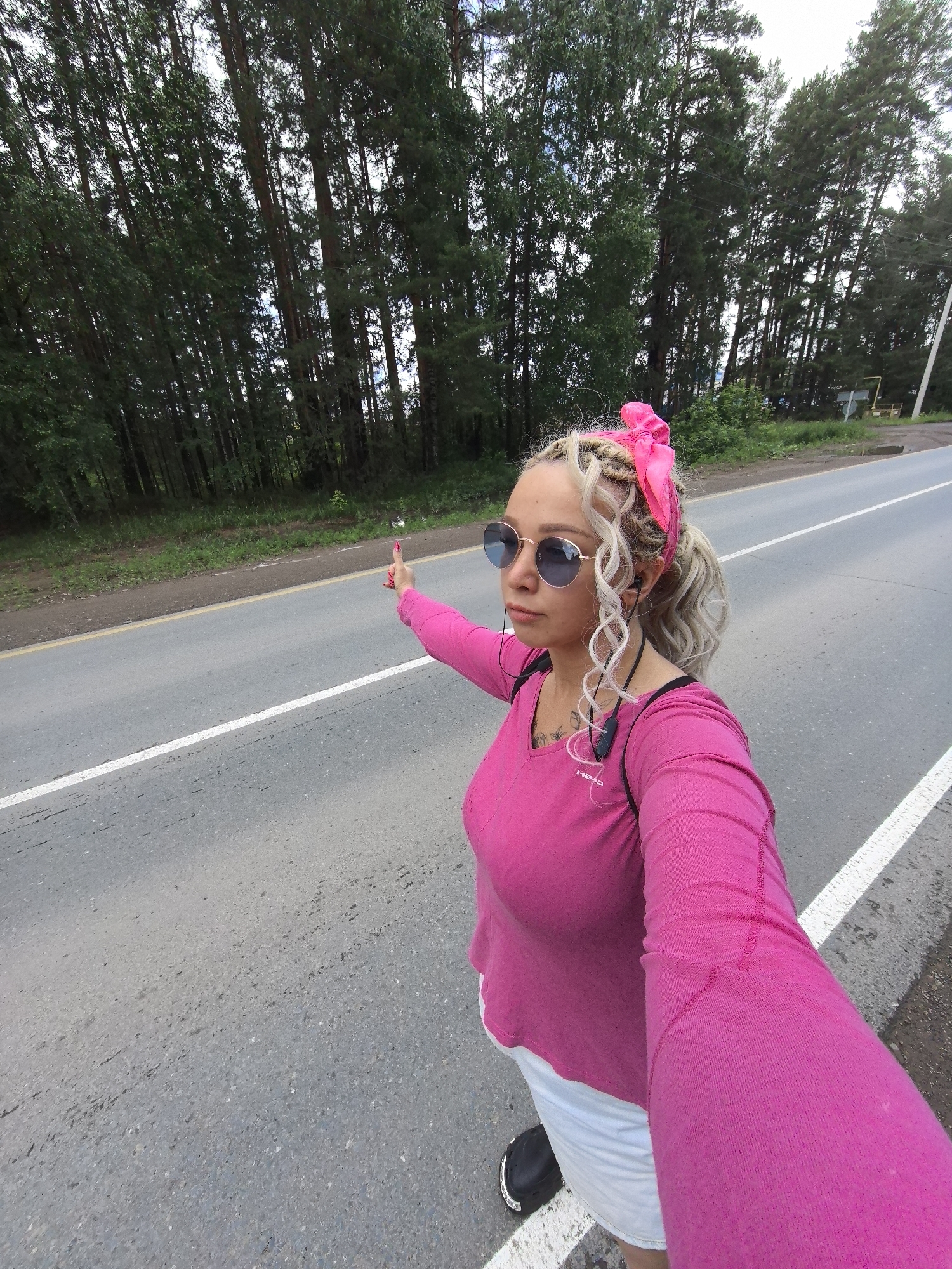 A woman with blonde braided hair and a pink bandana is standing on the side of a paved road, with trees lining the background. She is wearing a bright pink long-sleeved shirt, white shorts, black shoes, and round sunglasses. Her left arm is extended, and her thumb is raised, indicating she may be hitchhiking. She is also wearing black earbuds. The road has white lines marking the lanes and a yellow line along the shoulder. The environment appears to be a rural or suburban area in Noviy, Russia, given the dense tree line and the typical appearance of a secondary road.