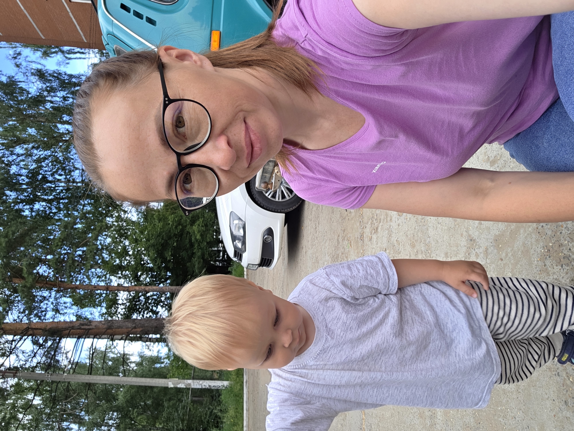 A medium shot selfie captures a woman with light brown hair, wearing black-rimmed glasses and a purple t-shirt, looking at the camera. To her left, a young child with blond hair, wearing a light gray t-shirt and striped pants, looks down. They are outdoors in what appears to be an urban or suburban environment. In the background, a white car is parked, and a teal vehicle is visible to the right. Trees and a brick building are also present. The ground is paved with dirt and gravel. The lighting suggests it is daytime. This scene is likely in Chaikovsky, Russia, given the context provided.