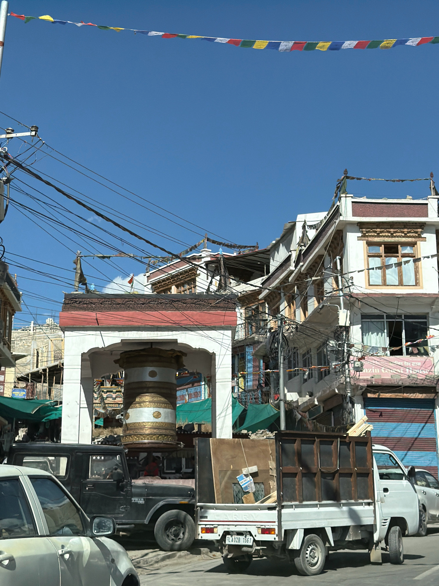 The image depicts a street scene in Leh, India, characterized by traditional architecture and a network of electrical wires overhead. In the foreground, a silver car is partially visible on the left, with its side mirror reflecting the surroundings. To its right, a black jeep is parked. Behind the jeep, a white pickup truck is loaded with lumber.
The background features multi-story buildings with ornate wooden window frames and balconies, a common architectural style in Leh. Prayer flags are strung across the street, adding a touch of color and cultural significance. In the center of the frame, a large, decorative cylindrical structure, possibly a prayer wheel, stands within an archway. A pink sign with text in a non-English script is visible on one of the buildings. The sky is clear and blue. The overall impression is one of a bustling urban environment with a strong cultural identity.