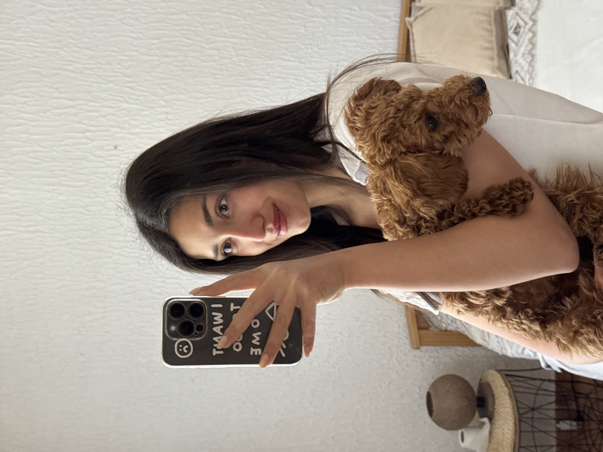 An indoor selfie captures a young woman holding a small, brown, curly-haired dog. The woman, with long dark hair and a light-colored top, looks directly at the camera with a gentle smile. She holds the dog in her left arm while her right hand, with light-colored manicured nails, holds up a smartphone. The phone's black case features white text that, when read correctly (as it appears mirrored in the selfie), says "I AM WHAT I AM," along with a partially visible smiley face icon.
The dog, resembling a poodle or similar breed, is nestled against the woman, looking upwards and away from the camera. The background shows a plain white wall, and parts of a bed with light-colored bedding and a wooden headboard are visible. To the lower right, a nightstand or small table is partially seen, featuring a round, textured decorative object. The scene appears to be set during the daytime, likely in a bedroom or living area. The image suggests a calm and affectionate interaction between the woman and her pet in a generic indoor setting in Sutomore, Montenegro.