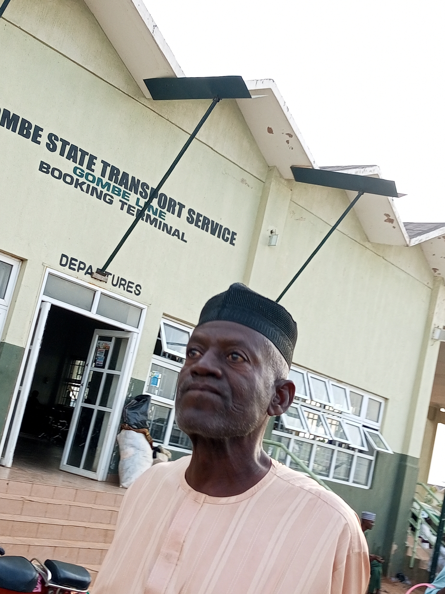 The image shows an outdoor scene at the Gombe State Transport Service Booking Terminal in Gombe, Nigeria. In the foreground, a man wearing a beige traditional outfit and a black cap is the primary subject. He is looking to his left, and his expression appears neutral. Behind him, the building of the transport terminal is visible. It is a light green structure with signage indicating "GOMBE STATE TRANSPORT SERVICE" and "GOMBE LINE BOOKING TERMINAL". Another sign reads "DEPARTURES" above a set of glass doors leading into the building. Several windows are visible on the building, some of which are open. Outside the entrance, there are steps leading up to the doors, and a motorcycle is partially visible in the lower left corner of the image. There are also some large white sacks, possibly luggage or goods, placed near the entrance. In the background, other people can be seen, including a person in a green outfit to the right and another figure inside the building. The sky is overcast, suggesting a cloudy day. The overall impression is of a functional transport hub.