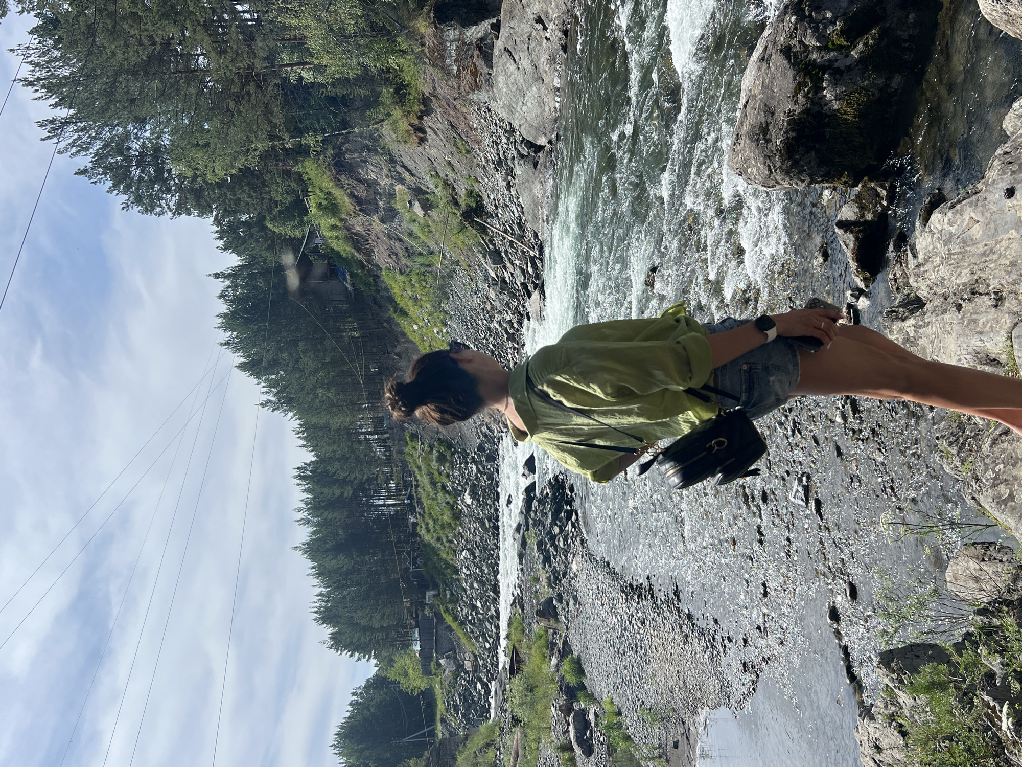 A woman stands on rocks overlooking a fast-flowing river in a mountainous, forested area. She is viewed from behind, wearing a light green, long-sleeved collared shirt, denim shorts, and a black backpack. The riverbed is rocky, with a gravelly shore to the left and larger boulders on the right. Dense green pine trees line the steep slopes of the mountains in the background. The sky is partly cloudy. There are some power lines visible in the upper portion of the image.