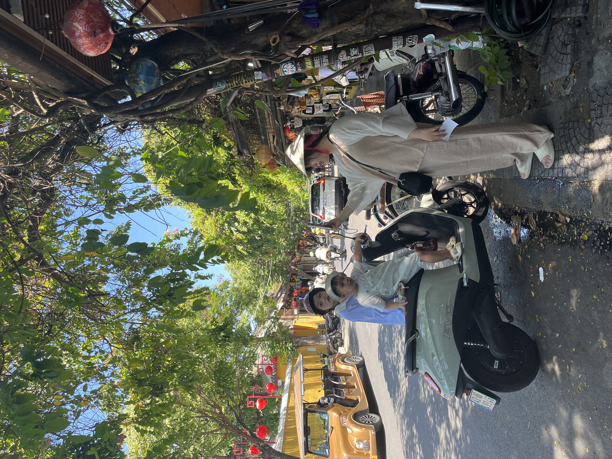 The image captures a vibrant street scene in Hội An, Vietnam, on a bright, sunny day. Lush green trees line both sides of the paved street, casting shadows and providing shade. The sky above is clear blue.
In the foreground to the right, a woman stands next to a light-colored scooter. She wears a white long-sleeved top, beige wide-leg pants, and white slide sandals. A traditional Vietnamese conical hat (nón lá) rests on her head, and she holds a small white piece of paper in her right hand. Behind her, another older motorcycle is parked.
On a light green scooter labeled "EVO", a young child in a striped outfit sits in front of an adult, who is wearing a light blue top and a helmet. The child looks towards the camera.
Further down the street, several other motorbikes are parked, and a distinctive gold-colored electric buggy, with "Hoi An" written on its side, is visible on the left. The street is lined with buildings featuring traditional architecture, adorned with numerous red and orange lanterns, characteristic of Hội An.
Overhead, entangled vines and branches are visible, along with decorative items resembling painted mask-like heads hanging from beams. Signs and stickers with Vietnamese text, including phone numbers like "0905 922714", are also visible on the overhead structure. The scene portrays a typical bustling day in a Vietnamese town, likely a popular tourist destination.