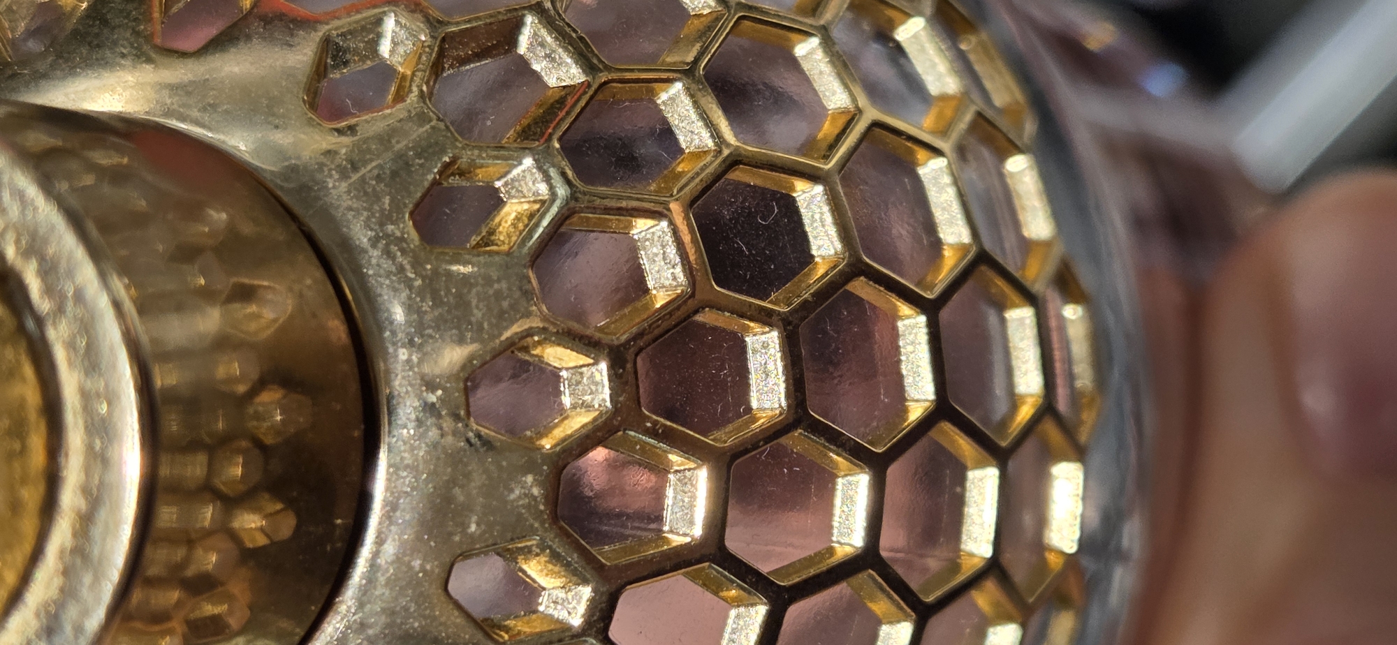 The image displays a close-up, macro view of a metallic, possibly gold-plated, object with a honeycomb-like pattern. The pattern consists of repeating hexagonal shapes with faceted interiors that reflect light. The metallic surface exhibits a textured, hammered appearance in some areas and a smoother, polished finish in others. The colors are predominantly gold, with darker, purplish-brown reflections within the hexagonal facets. The background is out of focus and appears to be a blurred environment, with hints of skin tone suggesting a hand is present, possibly holding or interacting with the object. The scene is static and devoid of any action. The image is a still photograph and provides no information regarding its location in Kehl, Germany, or any contextual details about people, subjects, objects, actions, interactions, or the environment.
