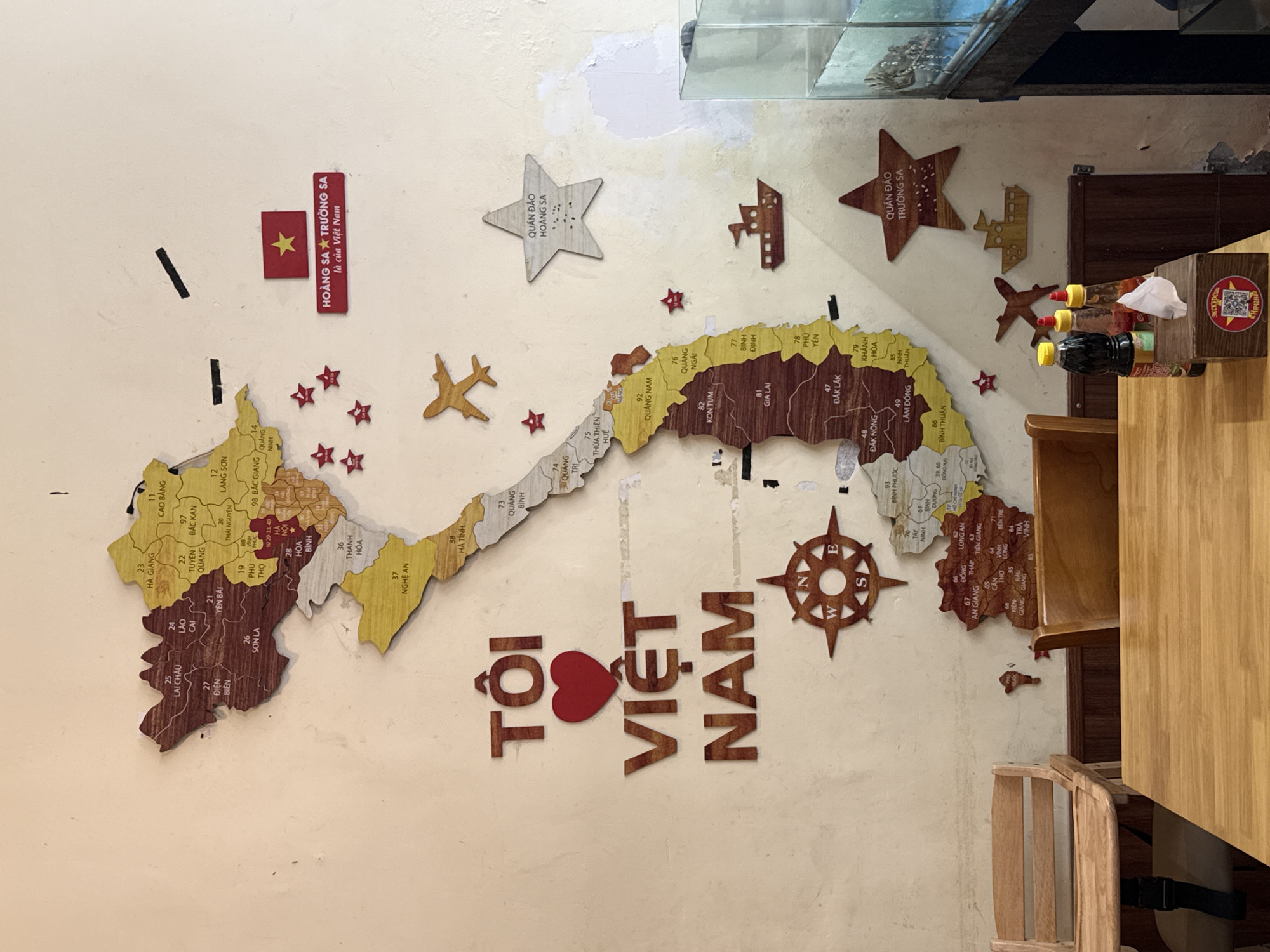 The image depicts a wall decorated with a large wooden map of Vietnam. The map is intricately cut and shows the country's provinces with numerical labels and names. Several wooden embellishments are attached to the wall around the map, including a Vietnamese flag, stars, an airplane, a ship, and a compass rose. A prominent wooden phrase "TÔI VIỆT NAM" (meaning "I love Vietnam") is centered on the wall.
In the foreground, a wooden table is visible, set with a wooden tissue box containing a packet of tissues. Two bottles of condiments, one dark and one red, are placed on the table. To the left of the table, a wooden high chair is positioned. To the right, a portion of a wooden chair is visible, and further to the right, a glass aquarium containing what appears to be fish or marine life is present. The wall itself is textured and shows signs of age and wear.
While the image is presented with a question about Nha Trang, Vietnam, there is no explicit visual information within the image that confirms its location as Nha Trang. The map itself is of Vietnam, and the decorations are country-specific. The context of the photograph, if it is indeed in Nha Trang, would be external to the visual content of the image.