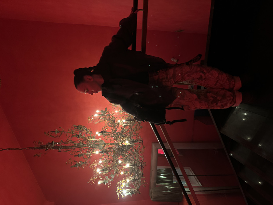 A person in a dark jacket and camouflage pants climbs a metal ladder within a red room. A large, elaborate chandelier hangs from the red ceiling. The room is dimly lit. A doorway and stairs are also visible. The scene suggests an unusual space with unique interior design.