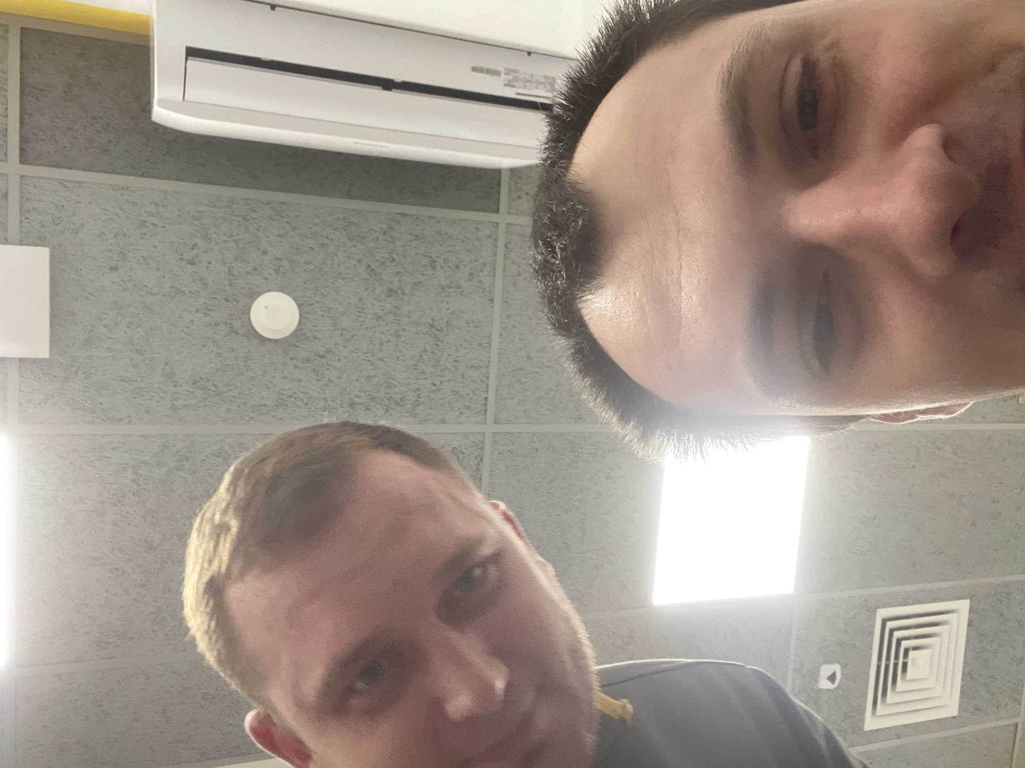 The image shows a selfie taken from a low angle, looking upwards towards the ceiling. Two individuals are partially visible. The person in the foreground is looking directly at the camera, with their face filling a significant portion of the frame. Their forehead, eyes, and nose are clearly visible. The second person is positioned to the left and slightly behind the first, with their head and part of their upper body visible. They are also looking towards the camera.
The environment appears to be indoors, likely an office or commercial space, as indicated by the suspended ceiling tiles with acoustic dampening material, a fluorescent light fixture, and an air vent on the ceiling. A white air conditioning unit is mounted on the right side of the wall. The lighting is bright, emanating from the ceiling fixture and possibly another light source below the frame. No specific identifiable landmarks for Boyarka, Ukraine, are present in the image.