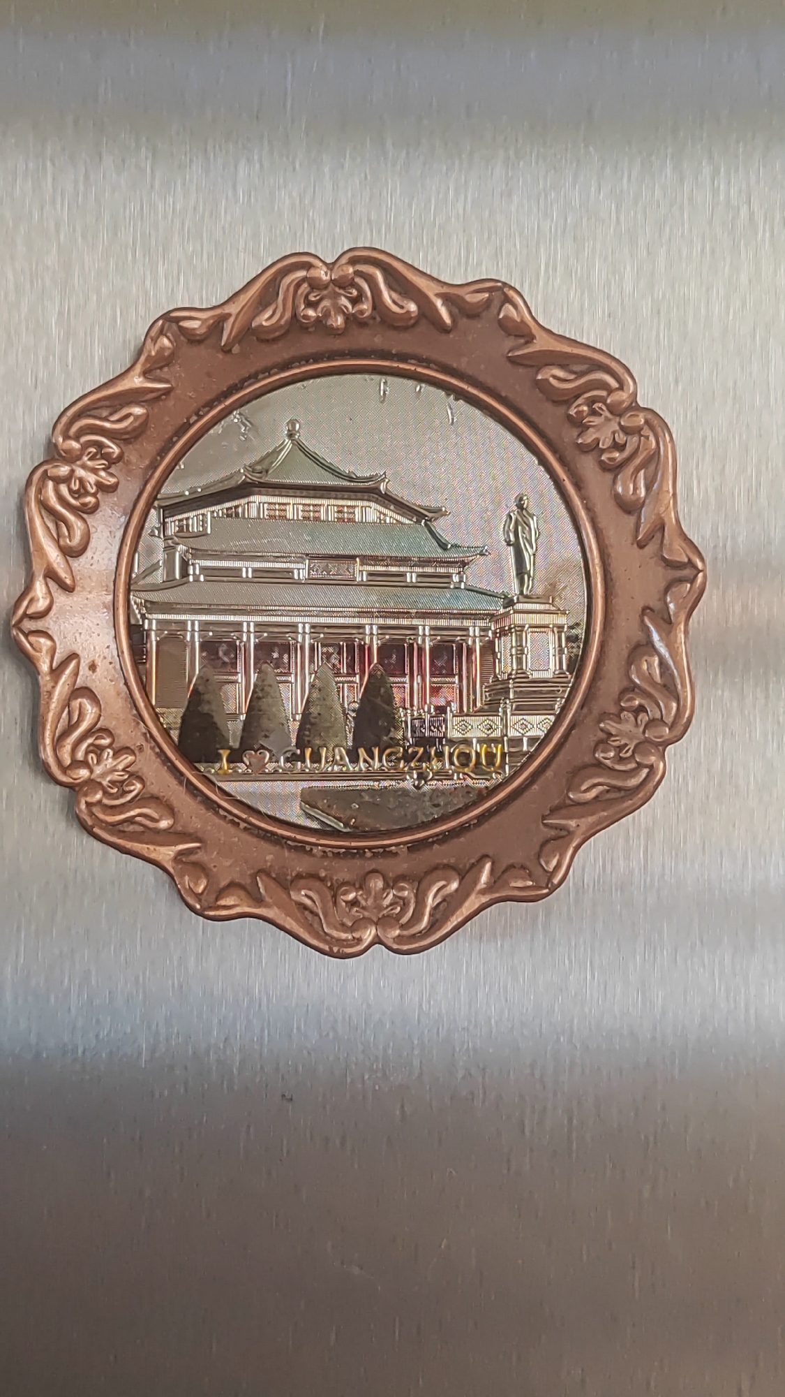 The image shows a souvenir plate with an embossed image of the Sun Yat-sen Memorial Hall in Guangzhou, China. The plate is round with an ornate, decorative border. The central image depicts the hall with its distinctive roof and colonnades, a statue of a man in a suit, and several cone-shaped trees in the foreground. Text below the trees reads "I <3 GUANGZHOU". The souvenir plate is attached to a brushed metal surface, likely a refrigerator or similar appliance. There is no indication of the city of Sochi, Russia in the image.