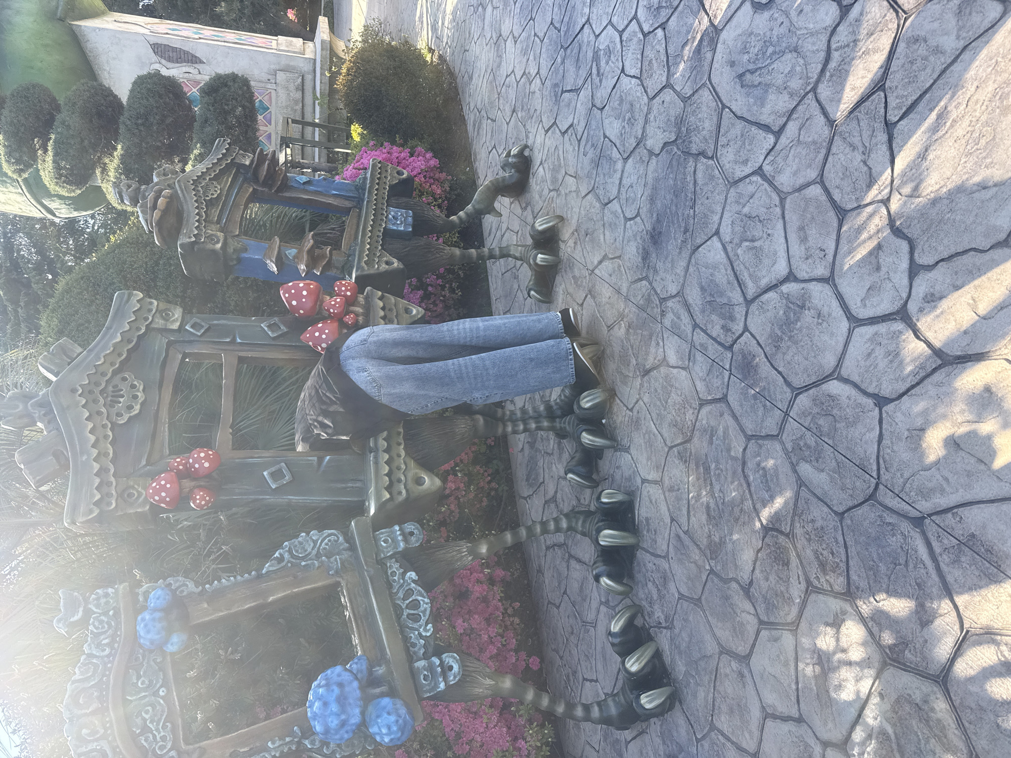 The image captures an outdoor scene at what appears to be a whimsical themed park or garden in Sochi, Russia. In the foreground, a person is partially visible from the waist down, wearing blue jeans and dark footwear, standing on a grey, irregularly patterned paved surface.
The central focus of the image is an array of fantastical, intricately designed structures that resemble enchanted huts or creature-like dwellings. These structures feature multiple dark, claw-like legs, giving them an anthropomorphic or animalistic appearance, similar to the concept of Baba Yaga's hut on chicken legs. They are adorned with elaborate carvings and painted details, including prominent red mushrooms with white polka dots and clusters of blue, berry-like decorations. The structures themselves appear to be made of wood or a similar material, featuring ornate frames, decorative rooflines, and a rich, earthy color palette with hints of blue and green.
Surrounding these structures are various green bushes and bright pink flowering plants, adding to the vibrant and magical atmosphere. In the background, more greenery and hints of other architectural elements suggest a larger, extensive themed area.
The scene is brightly lit by natural sunlight, casting long, distinct shadows across the paved ground, indicating either a morning or late afternoon setting. The clear lighting suggests fair weather. No text is visible in the image.