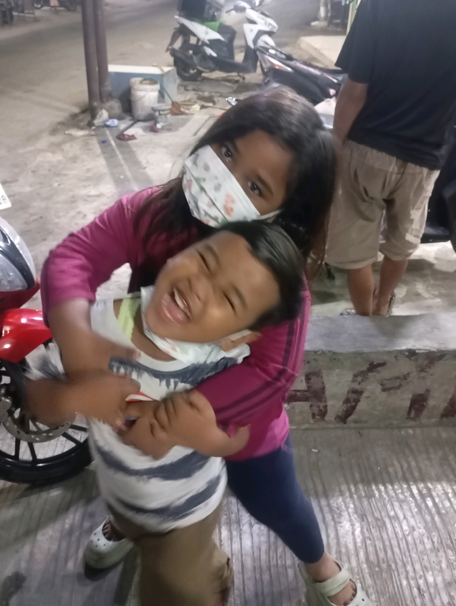 In Babelan, Indonesia, a young girl wearing a floral-patterned face mask hugs another child from behind. The girl in front is laughing, with her eyes closed and mouth wide open. She is wearing a striped t-shirt and what appears to be a light green undershirt. The girl hugging her is wearing a pink long-sleeved shirt. Behind them, a person in a black t-shirt and khaki shorts is visible, along with a red motorcycle to the left. The ground is paved with concrete, and there are some cylindrical objects and a white bucket in the background, suggesting an outdoor or semi-outdoor setting, possibly near a street or service area.
