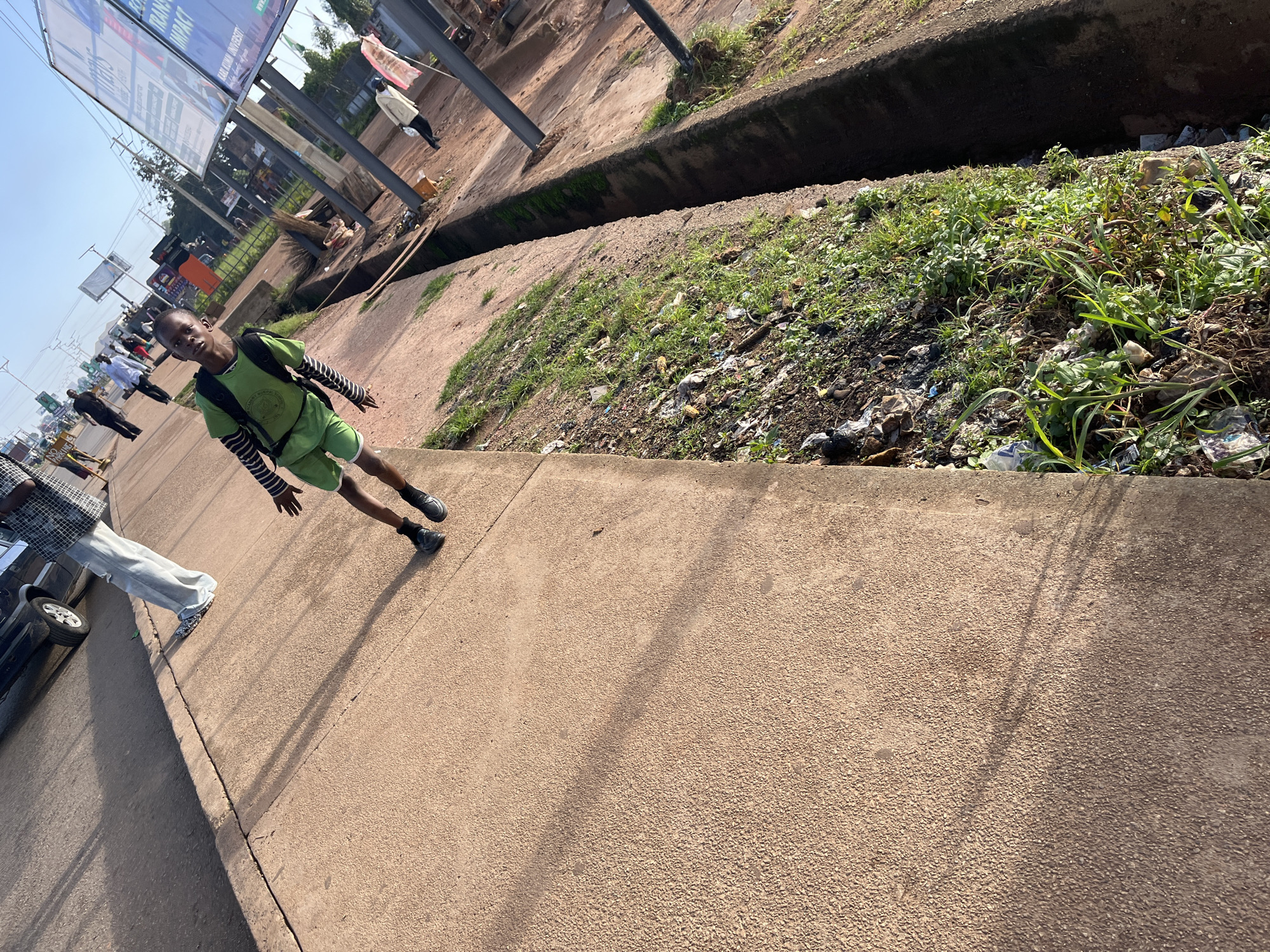 A young boy wearing a green shirt and shorts, and a backpack, stands on a paved sidewalk. He is looking at the camera. In the background, there are cars, people, billboards, and a drainage ditch filled with grass and debris. The scene appears to be a street in Gura Top, Nigeria. The lighting suggests it is daytime.