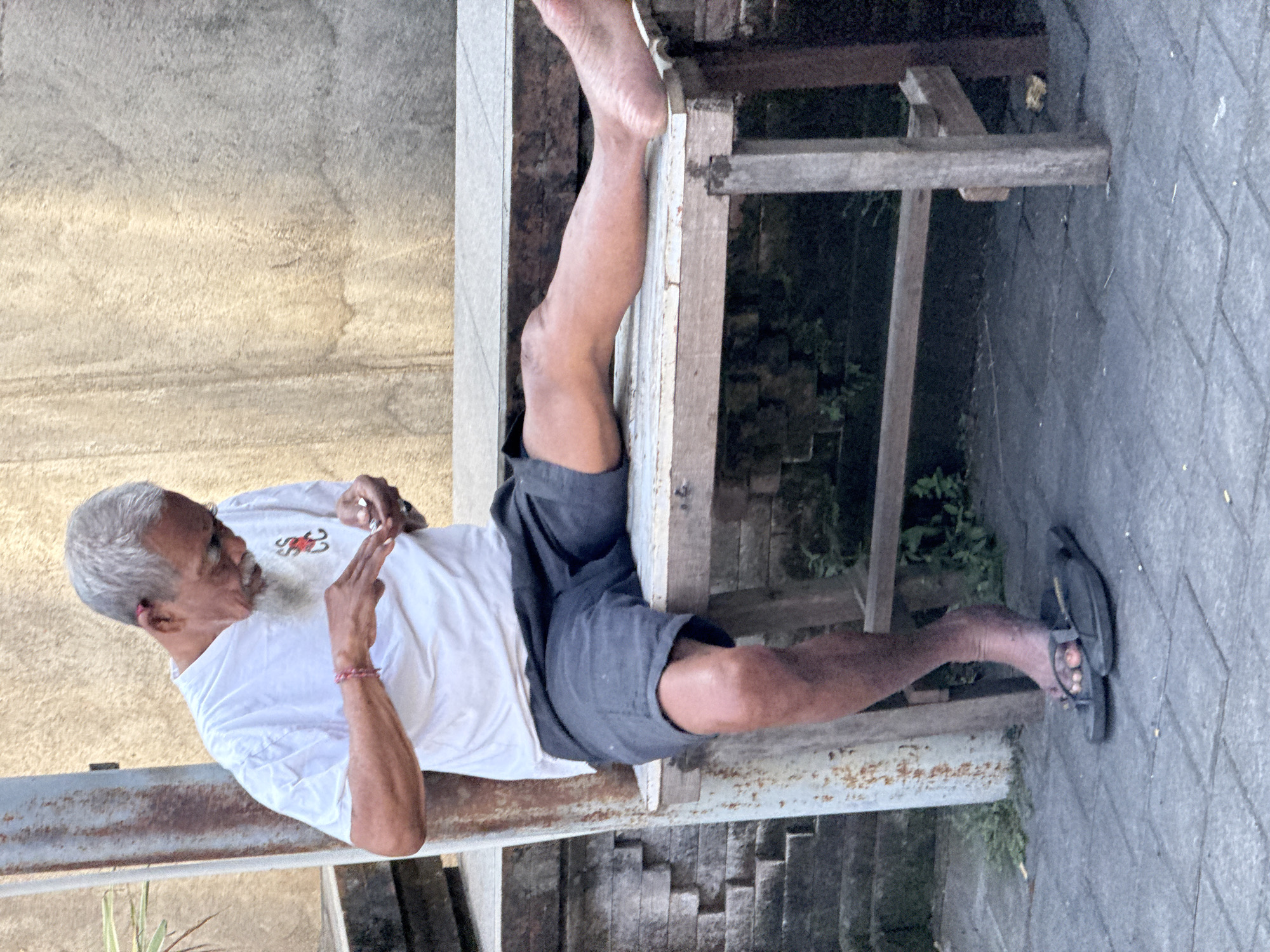 An elderly Asian man with gray hair and a beard is sitting on a wooden bench outdoors, possibly in a street or alley in Denpasar, Indonesia. He is wearing a white t-shirt with a red symbol on the left chest and dark shorts. His left leg is extended in front of him, resting on the bench, while his right leg is bent with his foot on the ground. He is holding something small in his hands and appears to be focused on it. The bench is weathered and appears to be made of unfinished wood, supported by simple wooden legs. Behind the bench is a textured wall, possibly concrete or stucco, with some water stains. To the left of the bench, a rusty metal pole stands vertically, and further to the left, there are stacked stone blocks. The ground is paved with gray rectangular tiles, and some greenery is visible beneath the bench. A pair of dark flip-flops are on the ground near the man's feet. The scene suggests a casual, everyday moment.