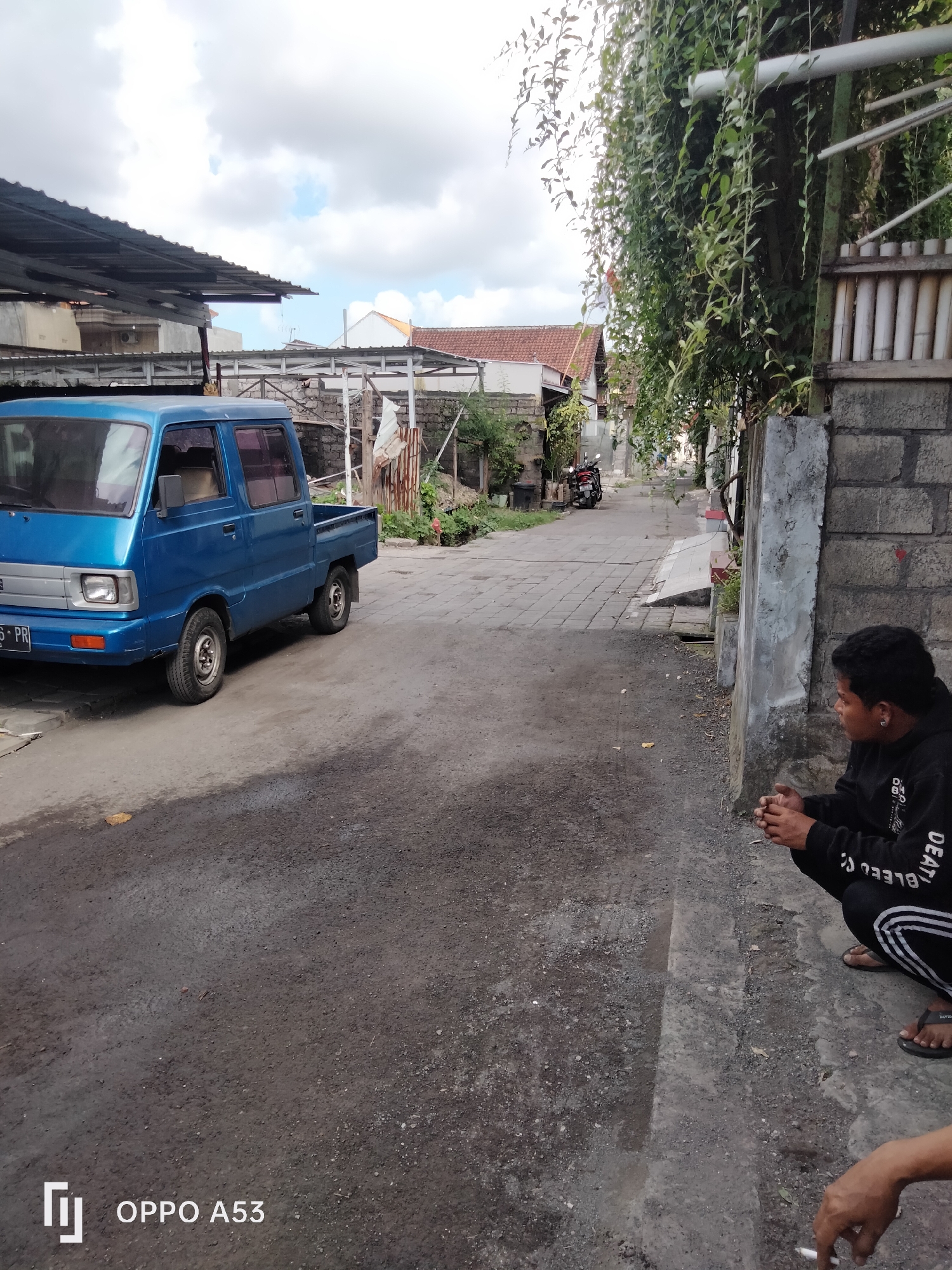 A person wearing a black hoodie and pants is crouching on a dirt and cobblestone street. A blue pickup truck is parked on the left side of the street. Buildings with red tile roofs line the street, and overgrown green vines hang from a fence on the right. The scene appears to be in a residential area of Denpasar, Indonesia, based on the architecture and vegetation.