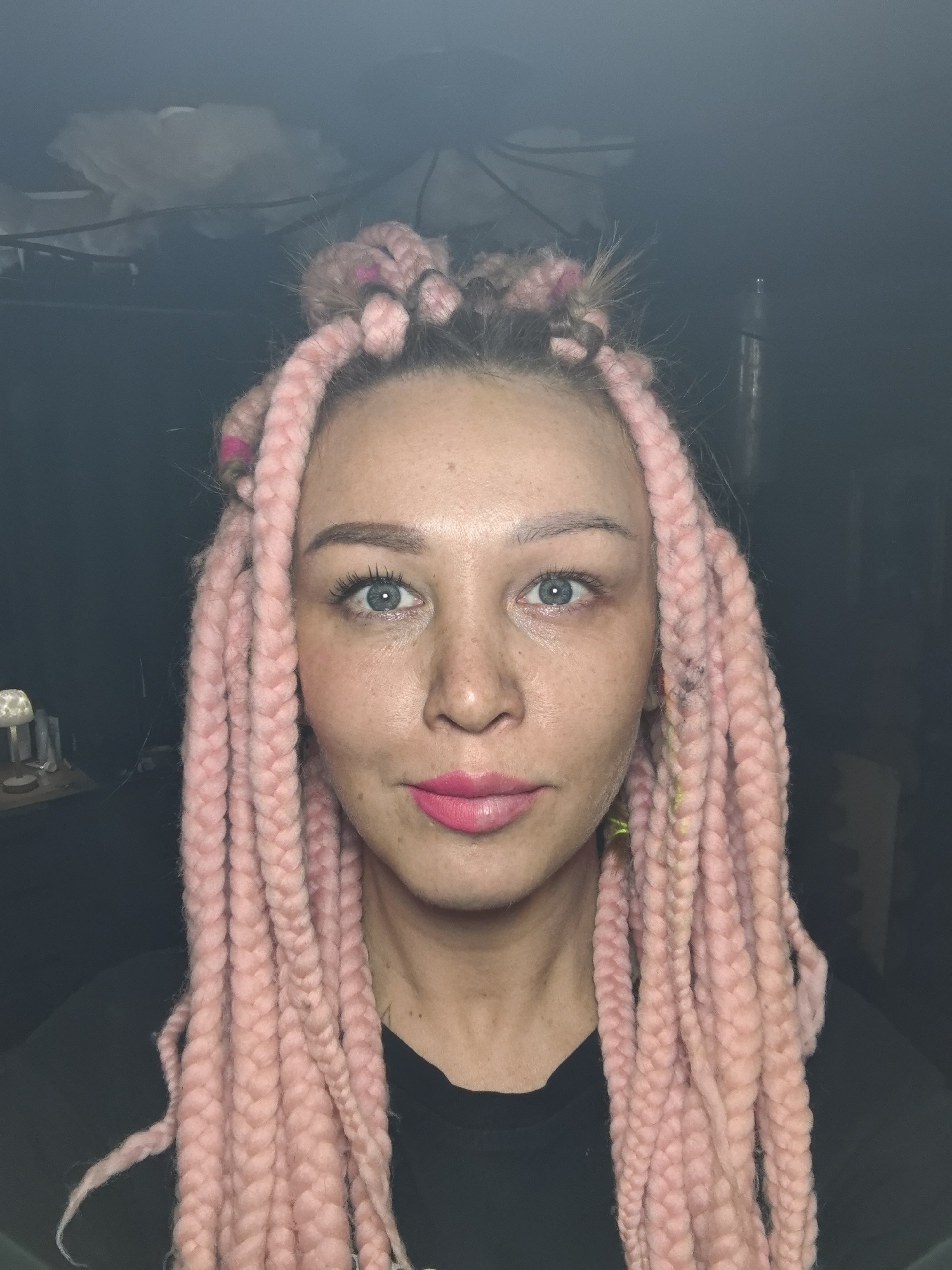 A close-up portrait captures a person with pink braided hair. The braids frame the face and are styled with a top knot. The person has fair skin, blue eyes, and is wearing pink lipstick. They are wearing a black shirt. The background is dark and indistinct, but a lamp and other items are dimly visible on the left. The location is the city of Noviy, Russia.