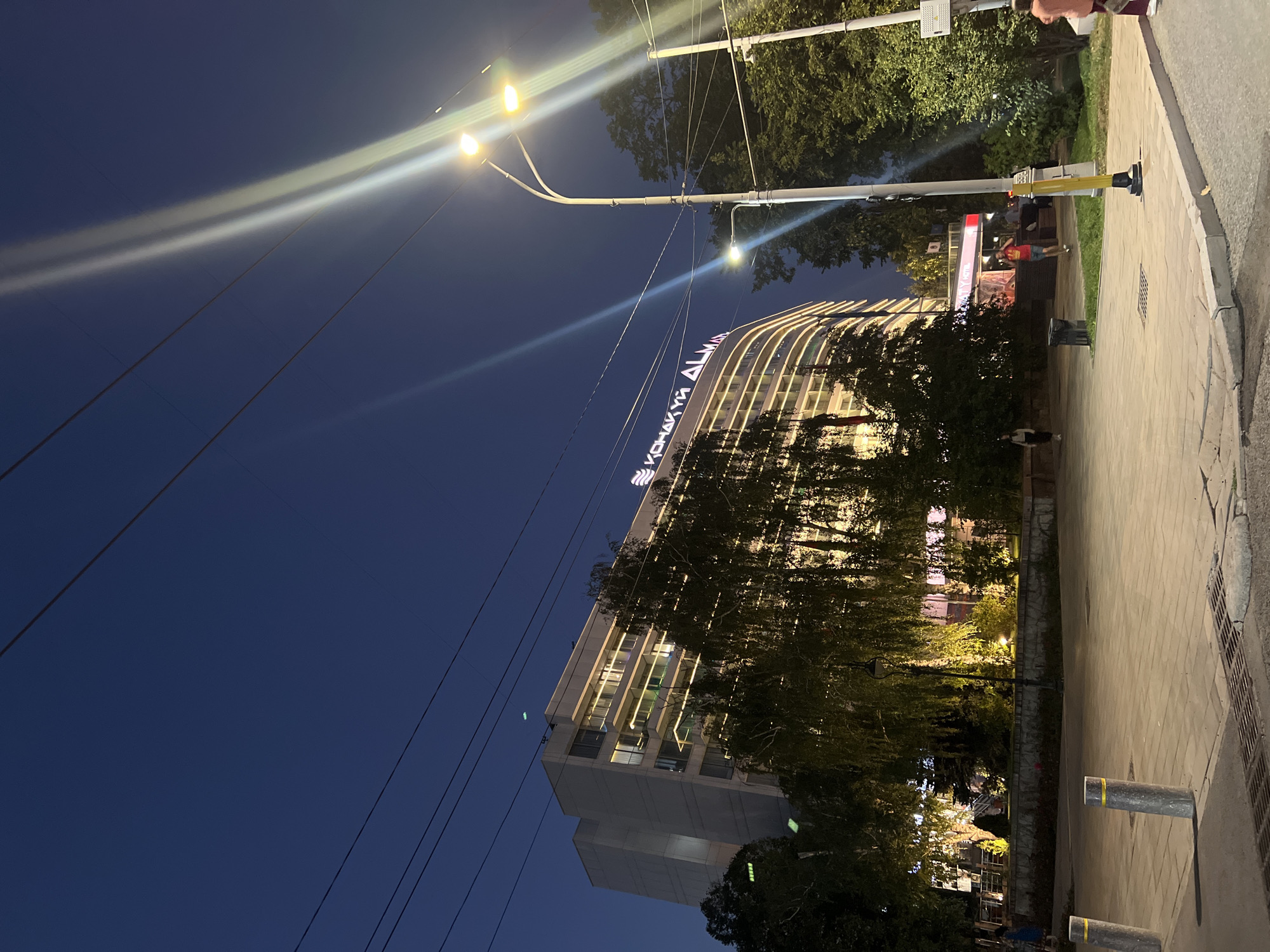 An exterior, low-angle, wide shot captures a scene at dusk in Almaty, Kazakhstan. The focal point is a large, modern, curved building with illuminated windows and decorative lights along its facade. Above the building, text in Cyrillic script is visible, reading "КОНАК УЙ ALMA" which translates to "Hotel Alma". To the right, a tall street lamp with multiple lights illuminates the area, casting long streaks of light across the image. Various trees, some with weeping branches, are situated in front of the building and in the foreground. The ground is paved with cobblestones, and concrete bollards are visible. Several indistinct figures are present: two women walking on the sidewalk, one man running on the sidewalk, and a woman with an orange top walking away from the camera on the far right. Power lines crisscross the dark blue sky, and the overall lighting suggests early evening.