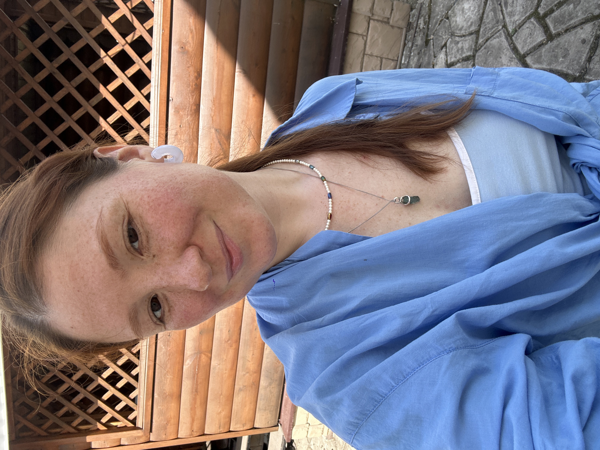 A portrait of a woman with auburn hair, freckled skin, and light eyes is centered in the frame. She is wearing a light blue collared shirt over a light blue tank top with a white trim. She is also wearing earrings and a necklace with a small green pendant. The woman is slightly smiling at the camera.
Behind the woman, there is a wooden structure, likely a fence or part of a building, with a lattice design at the top. To the right, there are wooden logs. The background suggests an outdoor setting, possibly a yard or garden.
The location is Nizhny Novgorod, Russia.