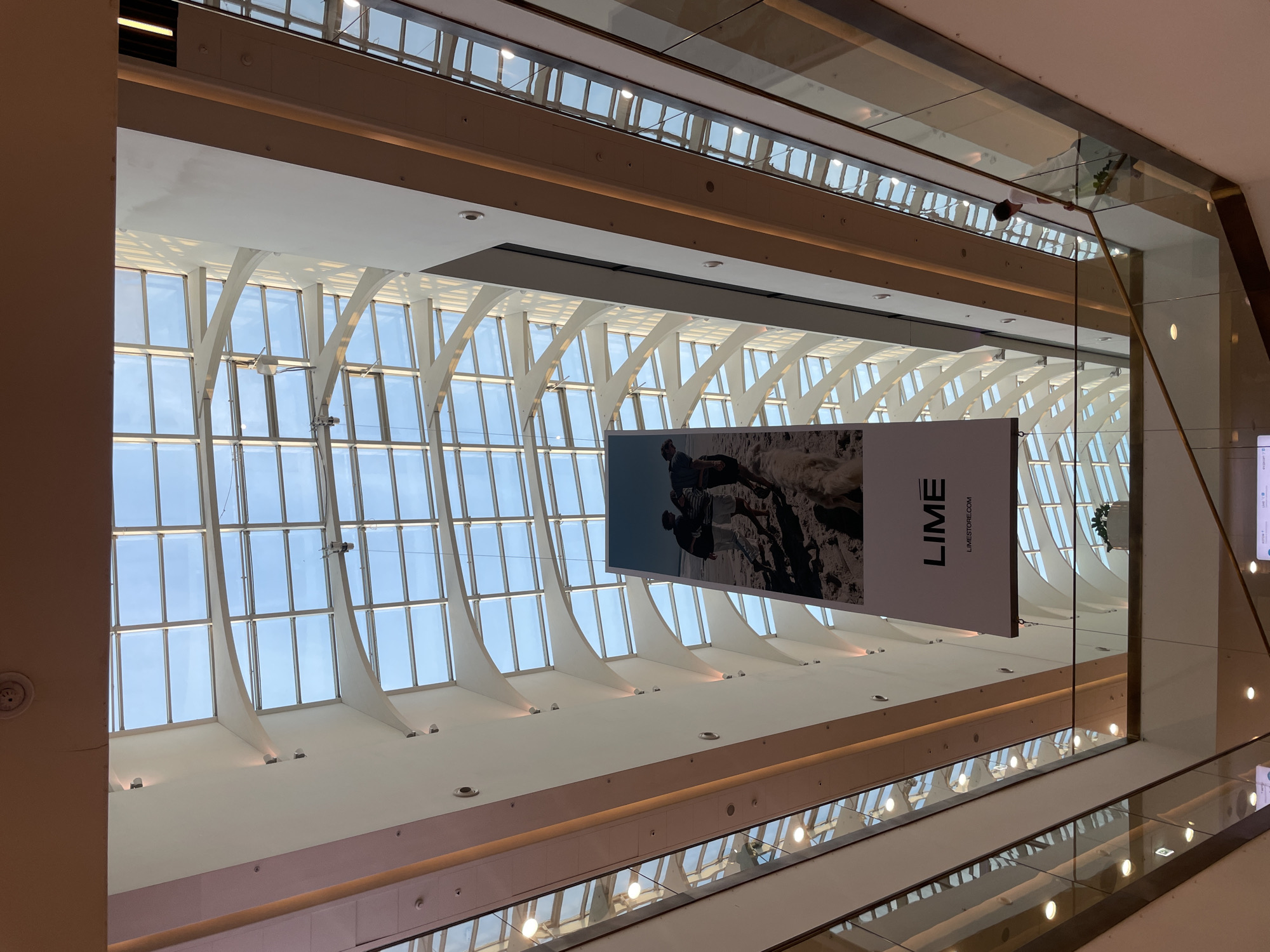Captured from a low angle, the image depicts the interior of a shopping mall in Saint Petersburg, Russia. The mall features a distinctive architectural design with a grid-like glass ceiling supported by white, curved support structures. This allows for ample natural light to filter into the space.
Hanging from the ceiling is a large rectangular banner with the word "LIME" printed prominently, accompanied by the website "LIMESTORE.COM." A black and white photograph adorns the banner, depicting two figures in a possible outdoor scene.
Below the banner, the mall's interior reveals multiple levels with glass railings. Lights are embedded in the ceiling, reflecting in the glass surfaces and creating patterns of light and shadow. The overall color scheme is dominated by whites, creams, and light browns, contributing to a sense of spaciousness and modernity.