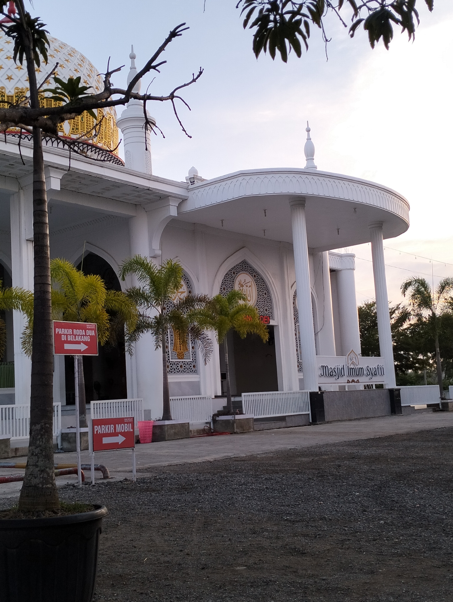 This is an eye-level, outdoor, medium shot of the Masjid Imum Syafi'i in Blangmetimu, Indonesia, taken during the daytime. The mosque is a white building with a prominent golden dome decorated with white stars. The mosque features a portico supported by white columns and intricate white latticework on its facade. Two red signs are visible: one indicating "PARKIR RODA DUA DI BELAKANG" (Motorcycle Parking in the Back) with a white arrow pointing right, and another reading "PARKIR MOBIL" (Car Parking) also with a white arrow pointing right. Several palm trees are present, with one in the foreground in a large black planter. The ground is covered in gravel. The sky is clear and light blue, with some tree branches visible in the upper portion of the image. The overall scene depicts a serene and well-maintained religious site.