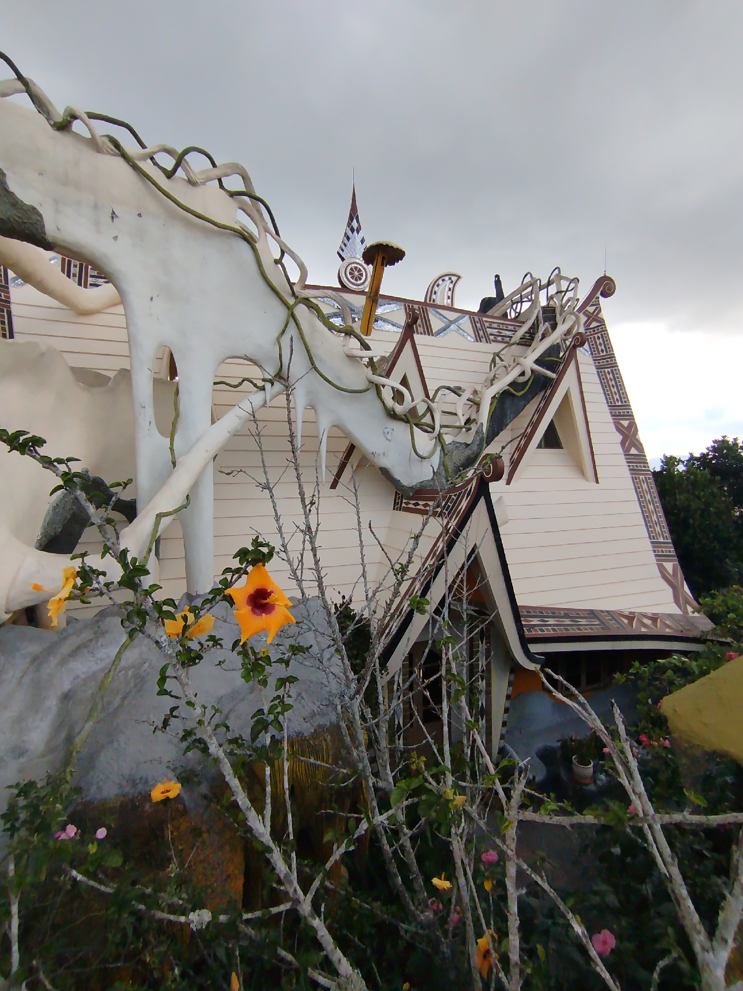 The image captures the Crazy House, also known as Hang Nga Guesthouse, located in Da Lat, Vietnam. The architectural style is unconventional, featuring elements that mimic natural forms such as trees, mushrooms, and animal figures. The main structure visible is a building with a traditional gabled roof, decorated with geometric patterns along the edges. A large, white, sculptural element resembling an animal or abstract form is integrated into the building's design. In the foreground, there's vegetation, including flowering plants with orange blossoms. The sky is overcast. There are no people visible in the frame.