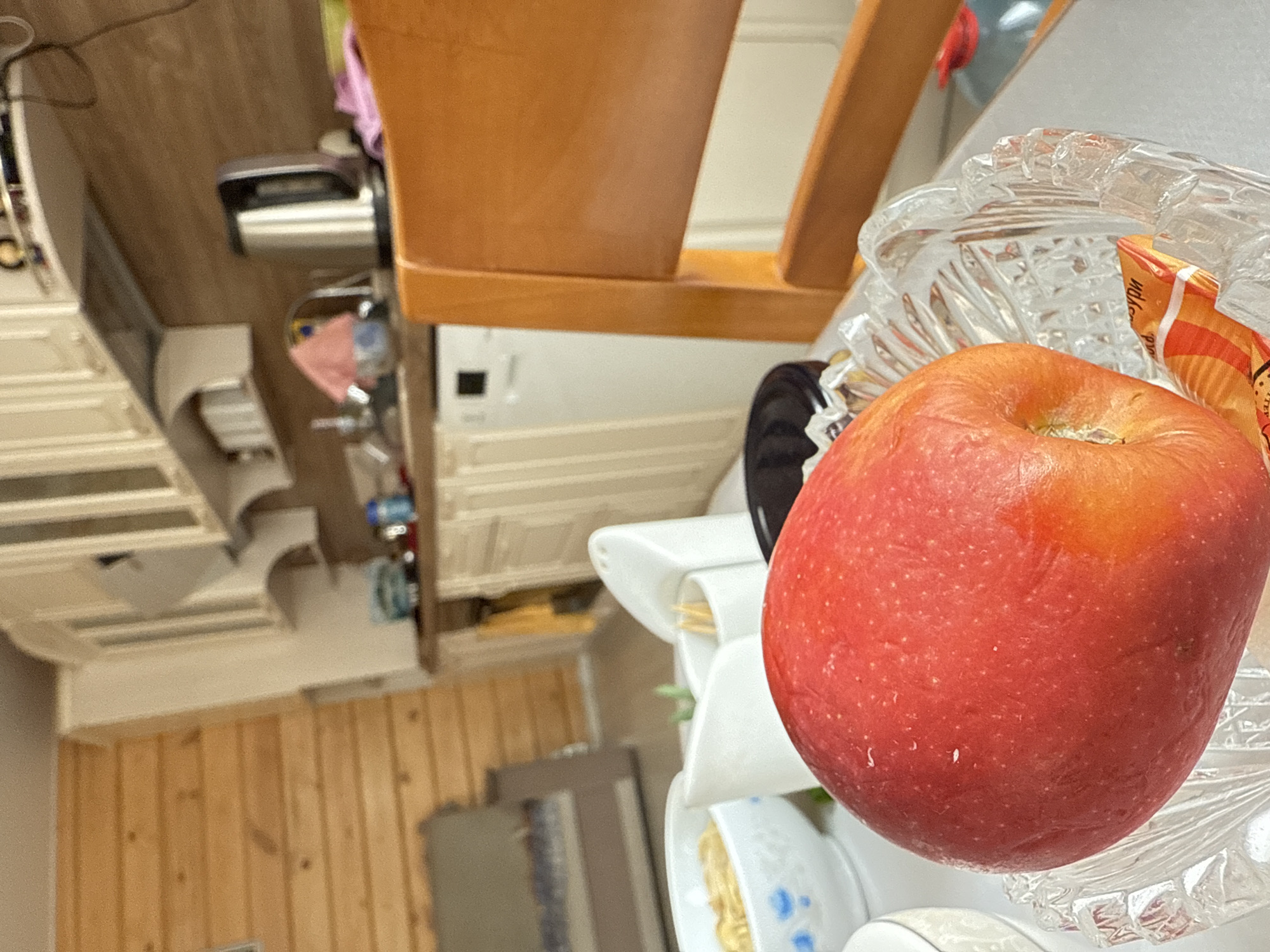 A close-up, tilted, and slightly rotated photograph depicts a red apple in a clear glass bowl, placed on top of a white, multi-tiered dish rack. The rack contains plates of food, including what appears to be noodles. The background features wooden floors and kitchen cabinetry, including a light-colored wooden cabinet and a white appliance, possibly a washing machine. Other kitchen accessories, such as a dish soap bottle and utensils, are vaguely visible. The location appears to be a kitchen interior, but the specific city and other details are not discernible from the image.