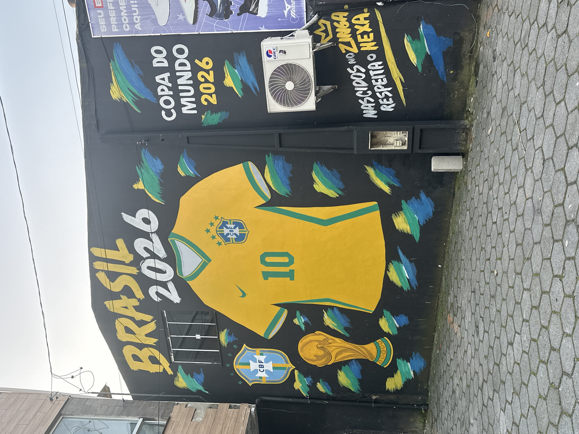 This is a medium shot, eye-level, outdoor photograph of a mural on a black wall. The mural depicts a large yellow soccer jersey with the number 10 and the Brazilian Football Confederation (CBF) logo. To the left of the jersey is a golden soccer trophy and the CBF crest. The words "BRASIL 2026" are painted in large yellow and white letters above the jersey, and "COPA DO MUNDO 2026" is painted to the right of it in white and yellow. The mural also features abstract brushstrokes in the colors of the Brazilian flag: green, yellow, and blue. Below the mural, graffiti reads "NASCIDOS NO ZINGA. RESPEITA O HEXA." An air conditioning unit is mounted on the wall to the right of the jersey. The ground in front of the wall is paved with hexagonal gray stones. The background shows a building with a tiled facade and a cloudy sky. A white sign with text in Portuguese is partially visible on the right side of the wall. This image was taken on Rua Doutor Arnaldo Quintão, in the city of Florianópolis, Brazil, on December 28, 2023.