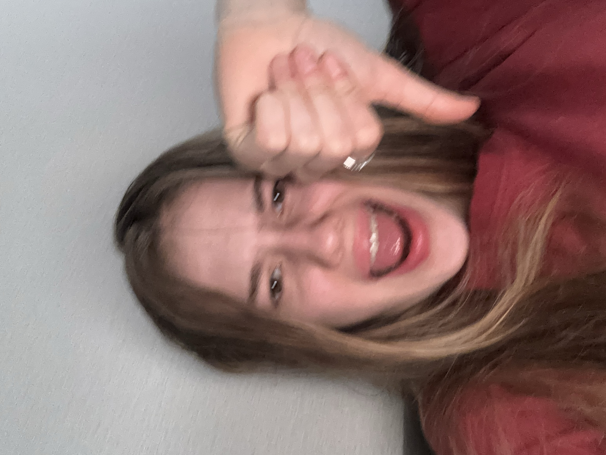 A young woman with long blonde hair and fair skin is shown in a close-up, blurry selfie. She has her mouth wide open in an expression of shock or dismay, with her teeth visible. Her eyes are wide and her eyebrows are furrowed. She is wearing a maroon-colored shirt. Her right hand is raised in the foreground, with her thumb pointing downwards in a "thumbs down" gesture. She is also wearing a ring on her right ring finger. The background appears to be a light-colored wall. The image does not provide any information about the city of Sinyavino, Russia, or any other geographical location.