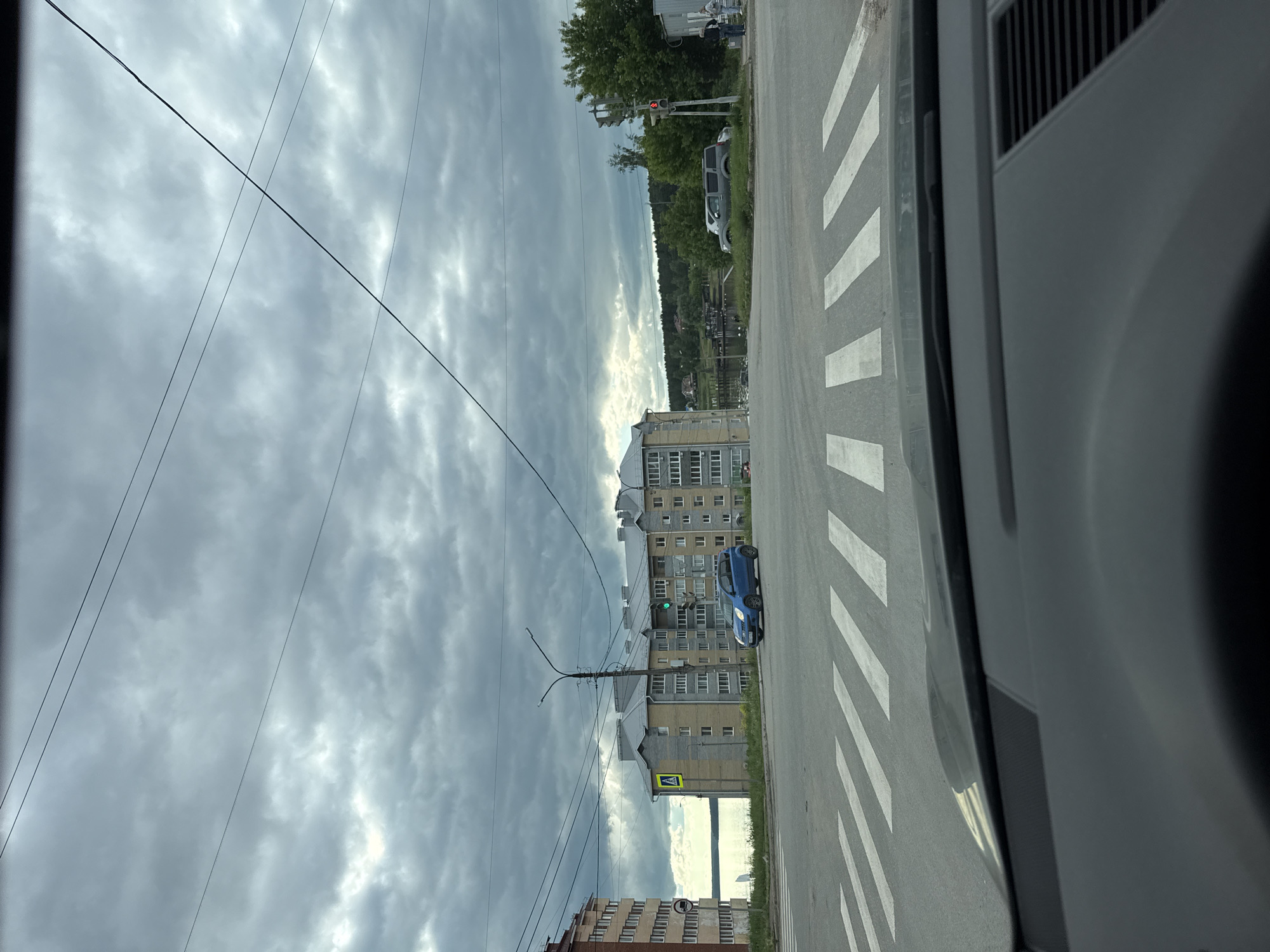The image, taken from inside a vehicle, captures an urban street scene on an overcast day in Votkinsk, Russia. The sky is dominated by dense, grey clouds, and numerous power lines traverse the upper portion of the frame.
In the foreground, a section of an asphalt road with a marked pedestrian crossing (zebra stripes) is prominent. The right side of the image shows parts of the vehicle's interior, including the dashboard and window frame.
Along the left side of the road, several multi-story residential buildings, consistent with post-Soviet architecture, line the street. A blue car is parked adjacent to one of these buildings. Near the curb, a yellow diamond-shaped road sign indicates an upcoming pedestrian crossing. A street lamp pole with intricate wiring stands on the sidewalk, and a traffic light showing a green signal is visible in the mid-ground.
Further down the street on the right, a grassy verge separates the road from a dense line of trees. Beyond the trees, more buildings and what appears to be a body of water or distant landscape are visible. A red traffic light can be seen on this side of the road, and a grey van is parked nearby. A few pedestrians are faintly visible walking in the distance.
The overall lighting is subdued due to the heavy cloud cover, suggesting daytime. The scene portrays a typical urban environment with no specific event unfolding, just everyday street activity. No explicit text is visible on signs, only symbolic representations.