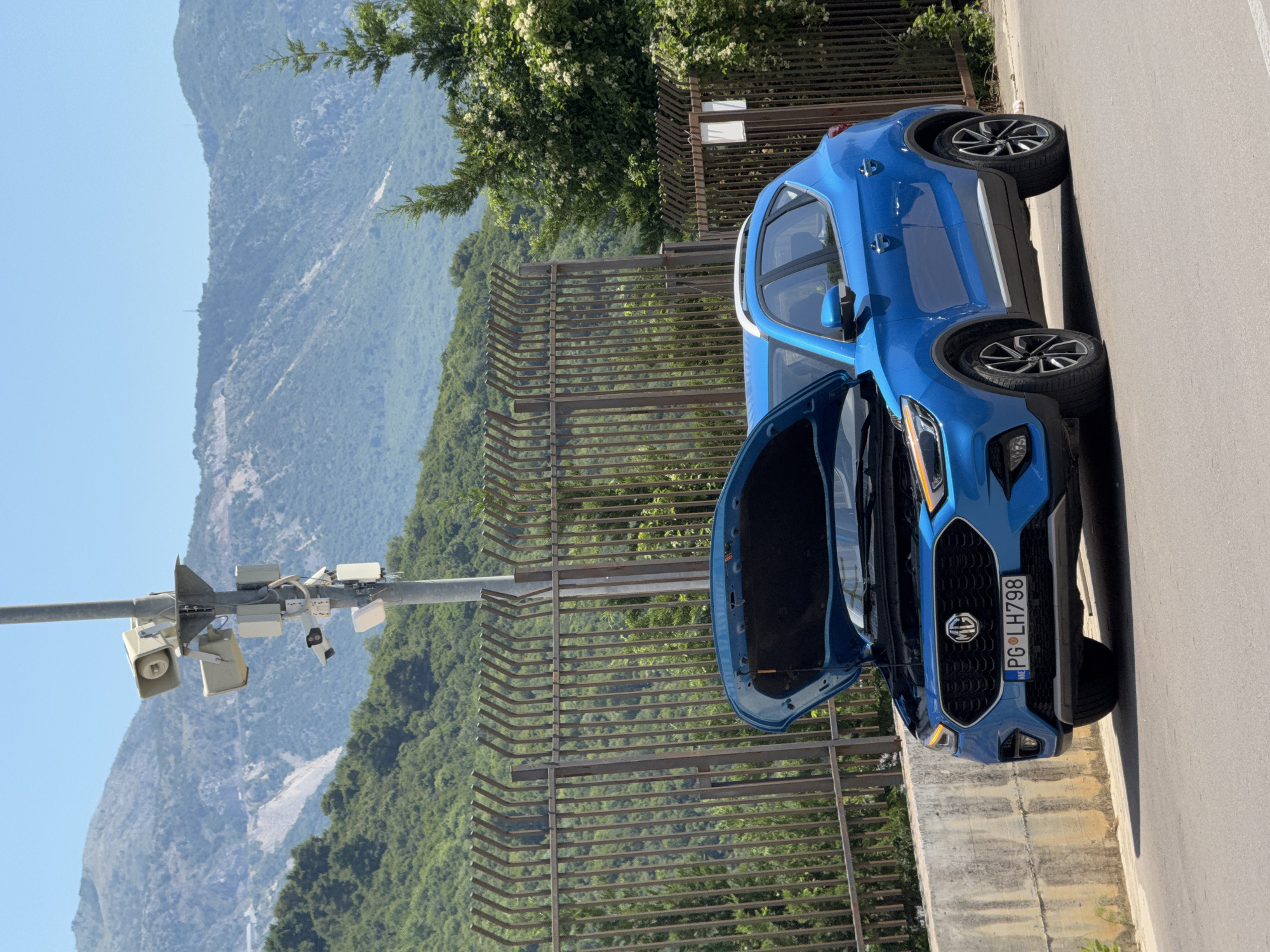 A blue MG ZS parked on a paved road, with its hood open, in Podostrog, Montenegro. The car, license plate PG LH798, is facing slightly towards the viewer, with the front grille and headlights visible. Behind the car, a tall metal fence separates the road from a hillside covered in dense green foliage. A pole with several cameras and speakers stands near the fence. In the background, a steep, rocky mountain range dominates the landscape, with patches of vegetation. The sky is clear and blue.