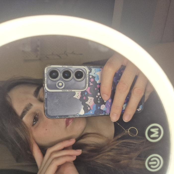 A mirror selfie captures a woman holding her phone. Her phone case is clear with cartoon cat and star designs. The phone has a multi-lens camera system. The woman has dark hair and light skin. Her face is partially obscured by her hand. The mirror is circular, possibly part of a vanity or makeup setup, and includes illumination elements around its perimeter and power buttons on the bottom.