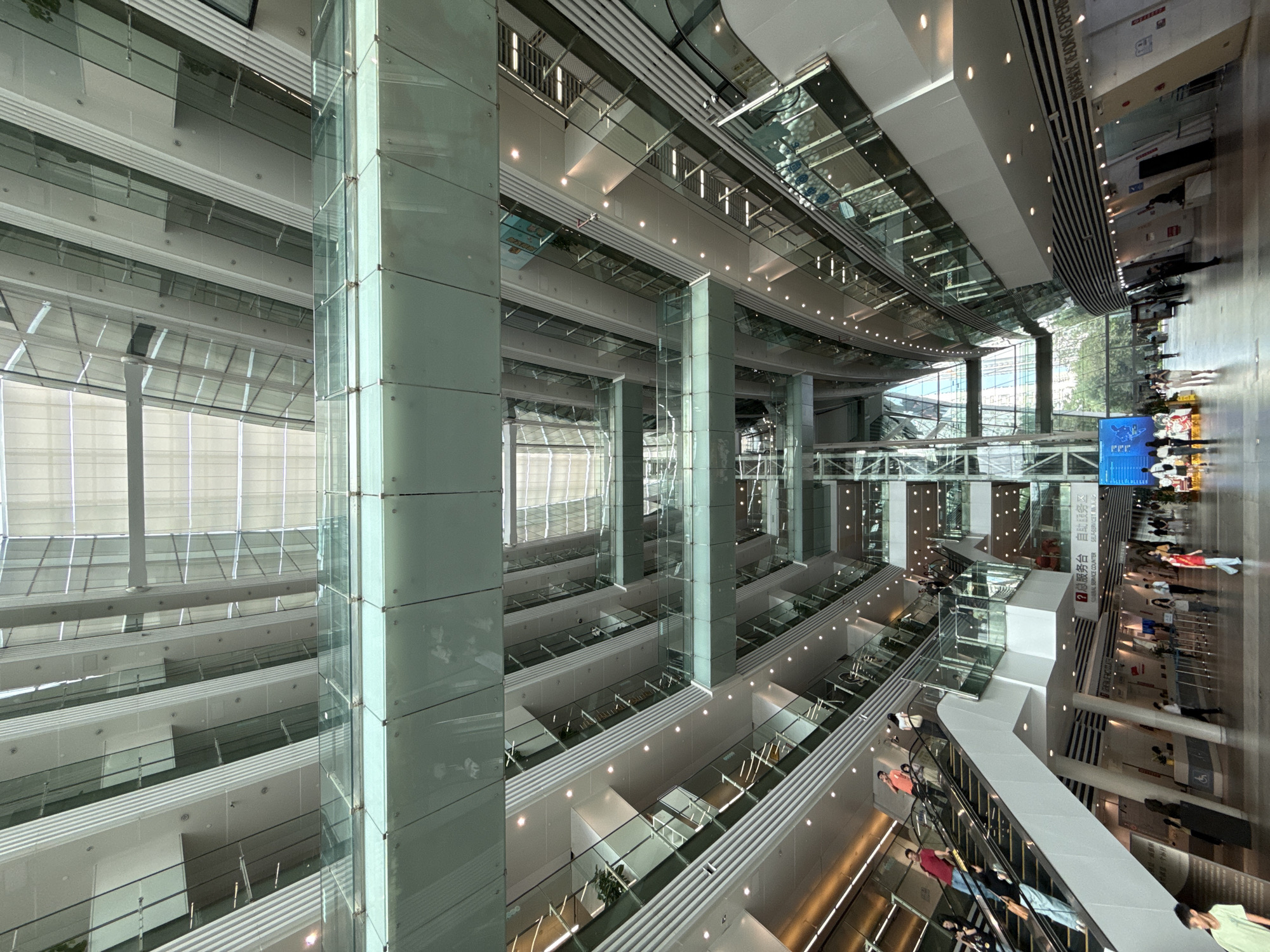 Here is a detailed description of the image:
**Environment:** The image captures the interior of a modern, multi-story building with a large central atrium. The design emphasizes glass and steel construction, with numerous levels visible. The architecture is characterized by clean lines, symmetrical arrangements, and an abundance of natural light.
**Objects:** Glass panels and supports are predominant, forming the railings for the various floors. There are rows of recessed lights illuminating the ceilings. In the far background, it appears that there are blinds on the windows.
**People:** The presence of people along the right edge of the image suggests the scale of the space. They appear to be walking or waiting in a queue.
**Actions/Interactions:** People are engaging in pedestrian traffic within the building.
**Location:** The image likely originates from a contemporary architectural design or a public space inside a modern building. The city of Unknown, Unknown is not identifiable within the image.