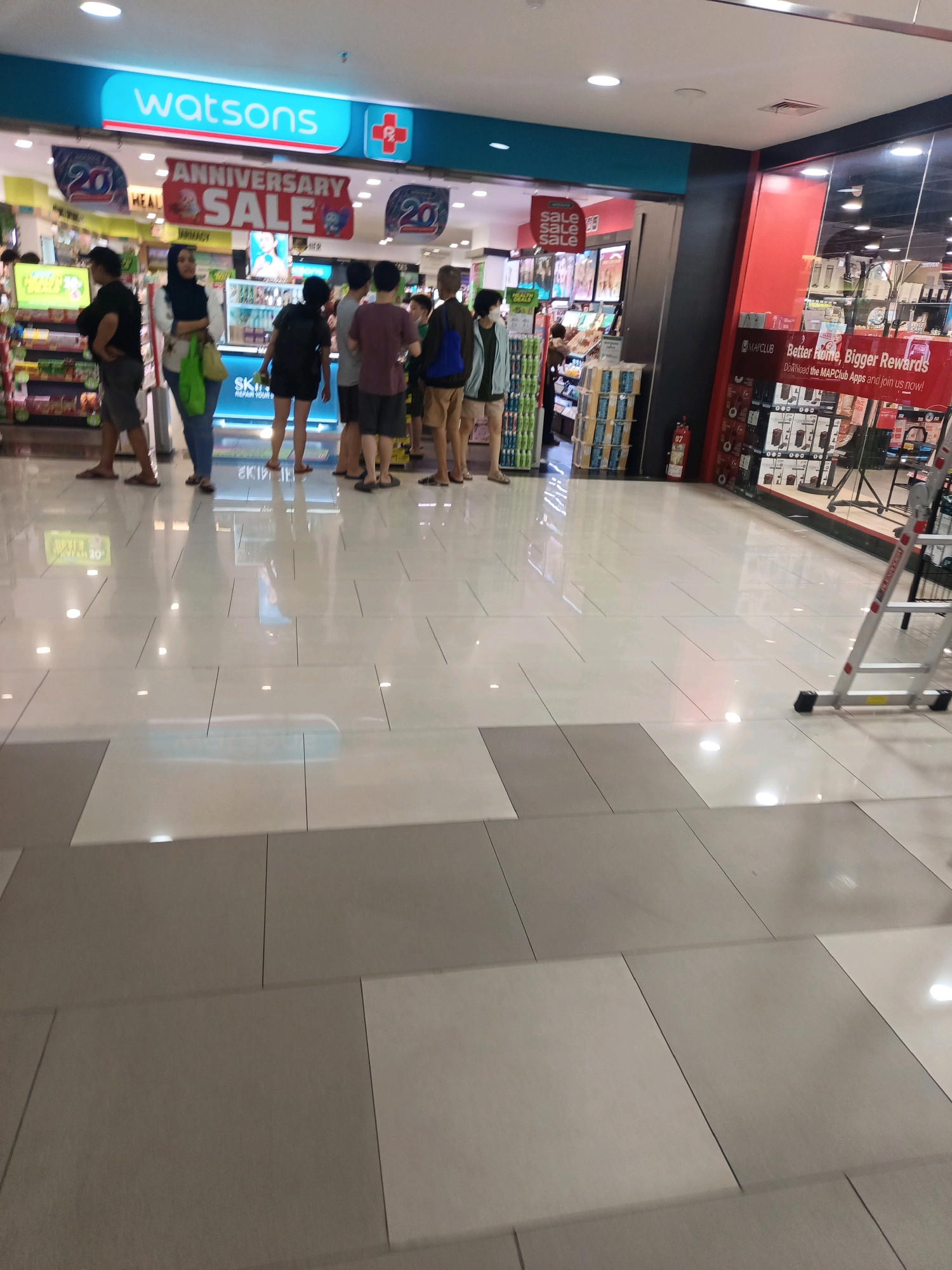 The image shows a Watsons store in North Jakarta, Indonesia, during an anniversary sale. In the foreground, several people are standing and looking at displays. A ladder is visible in the right side of the image, suggesting some sort of display setup or maintenance. The store appears to be well-stocked with various products. The floor is tiled with light-colored tiles, and the lighting is bright. The overall atmosphere seems to be a typical retail environment.