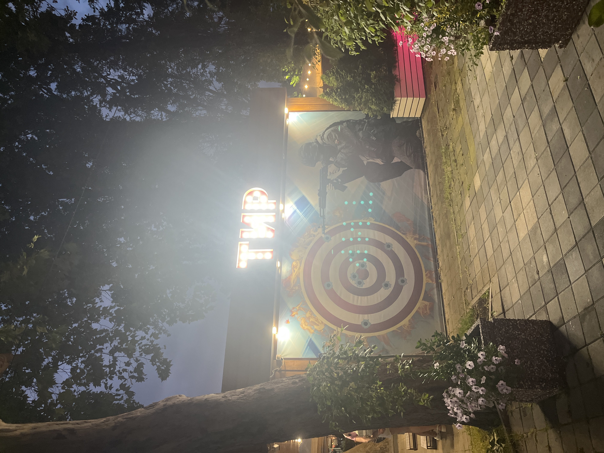 In Krasnodar, Russia, a shooting gallery attraction is depicted at dusk. A large, illuminated sign with Cyrillic lettering spelling "ТИР" (TIR) is prominently displayed above a mural. The mural features a circular target with bullet holes and a depiction of a soldier aiming a rifle. The scene is set outdoors, with paved pathways, planters containing flowers, and trees forming the surrounding environment. The lighting suggests it is evening, with artificial lights illuminating the attraction and the sky appearing dark blue.
