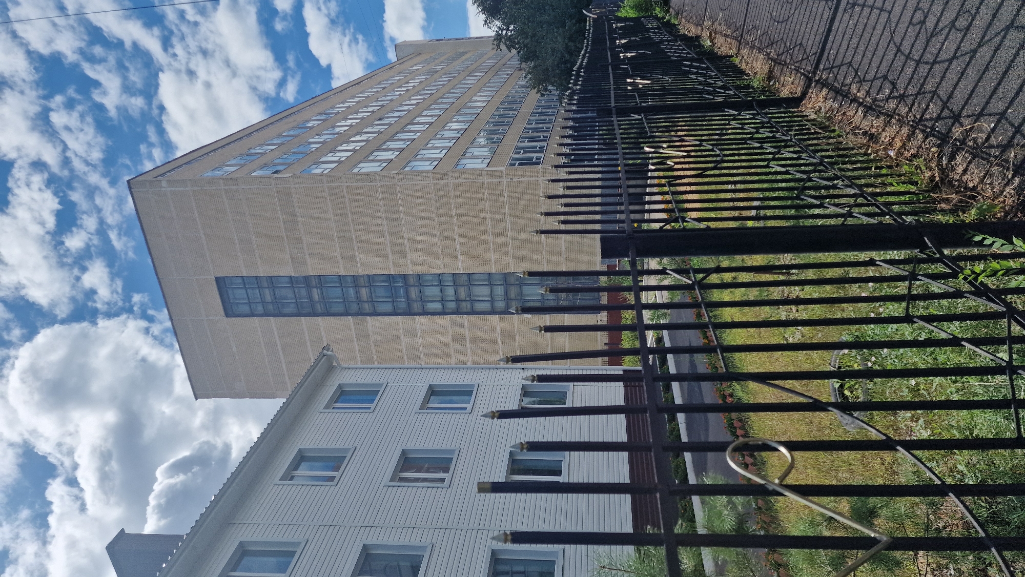 The image shows a long, light-yellow brick building with a tall, narrow vertical section of windows in the center, and rows of windows along its side. To the left of this building is a shorter, white-paneled building with multiple windows. A black metal fence with pointed tops runs along the foreground, separating a sidewalk from a grassy area with some plants. The sky is blue with scattered white clouds. The scene appears to be a street view of an urban area, likely part of a complex or campus. The context provided indicates this location is in Kostanay, Kazakhstan.