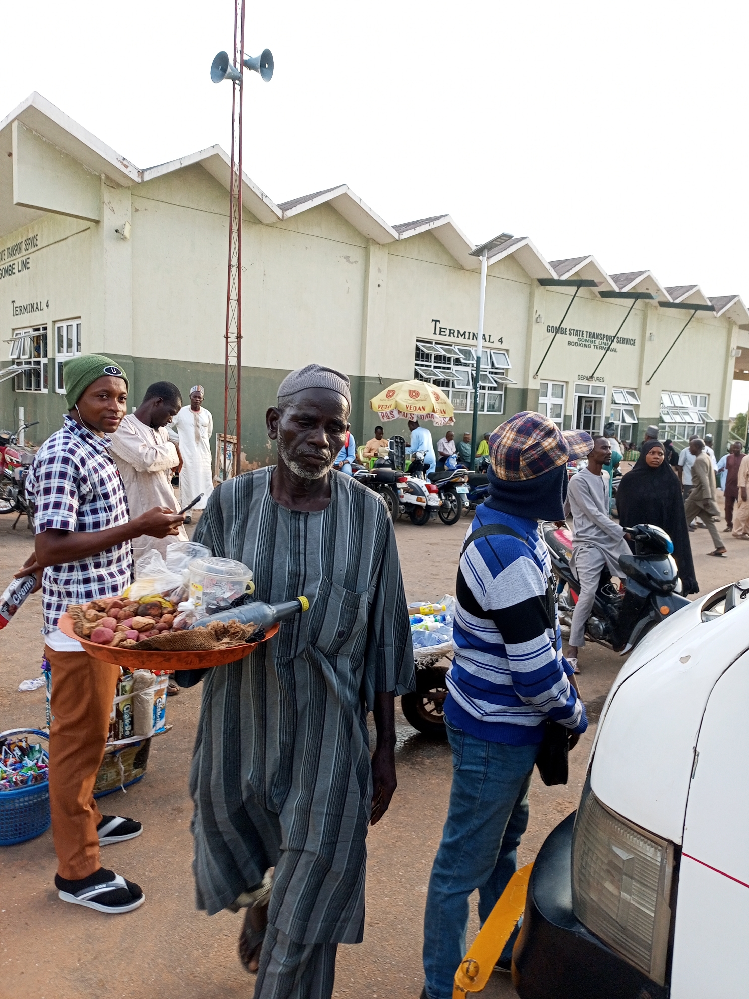 The image depicts a bustling outdoor scene at Terminal 4 of the Gombe State Transport Service, Gombe Line Booking Terminal in Gombe, Nigeria. The foreground is dominated by two men. The man on the left, wearing a green beanie and a plaid shirt, holds a smartphone and stands near a basket filled with colorful items. To his right, an older man in a striped robe carries a large tray laden with potatoes and other goods. Behind them, several individuals are visible, including a man in a white garment standing further back, and people milling about near motorcycles and a white vehicle. The terminal building, with its distinctive zigzag roofline, is visible in the background, marked with signage indicating "Terminal 4" and "Gombe State Transport Service." The overall atmosphere suggests a busy transportation hub with vendors and travelers present.